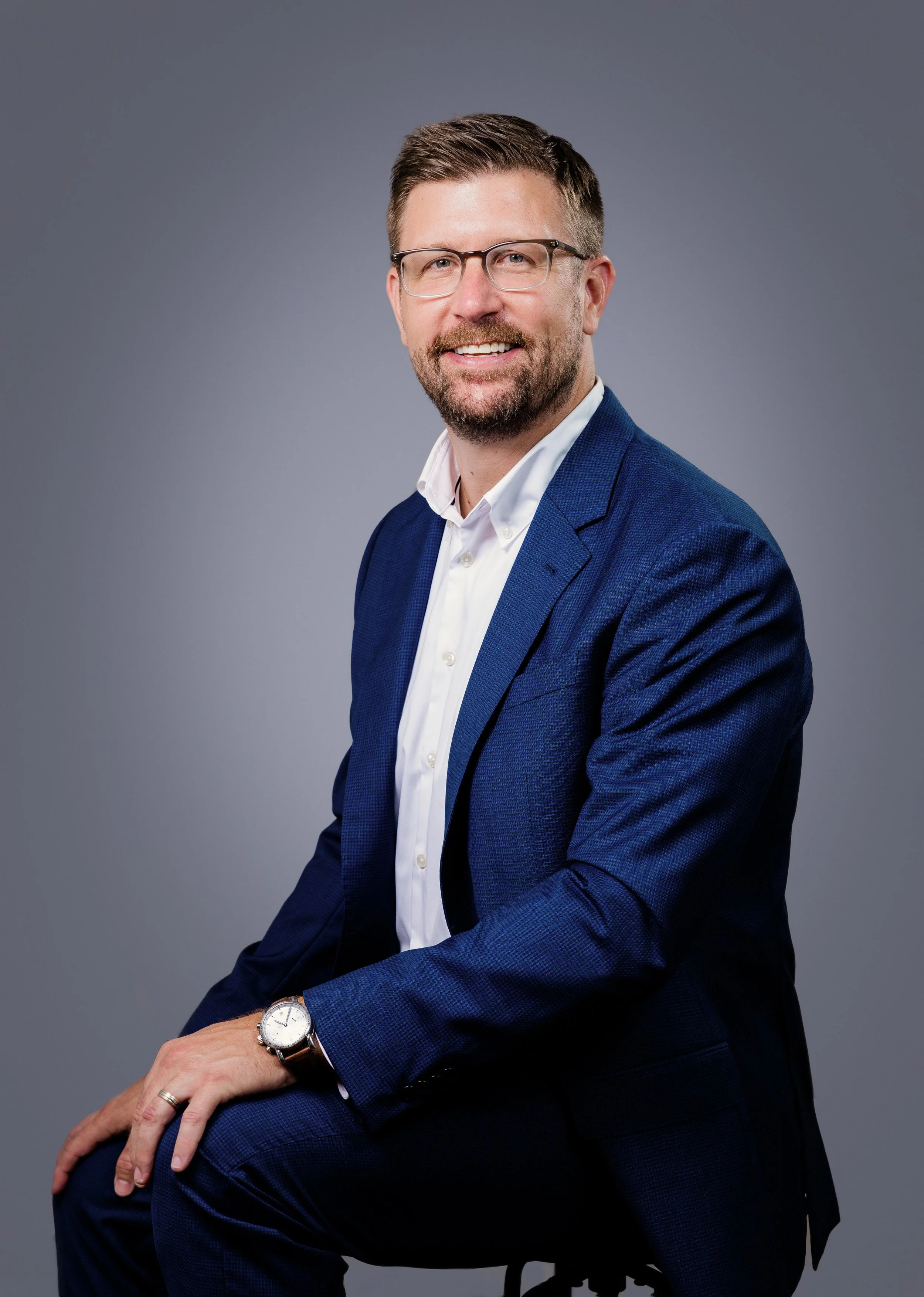 Portrait of a smiling man in a navy blue suit, white shirt, wearing glasses, with a watch on his left wrist, sitting on a stool against a gray background.