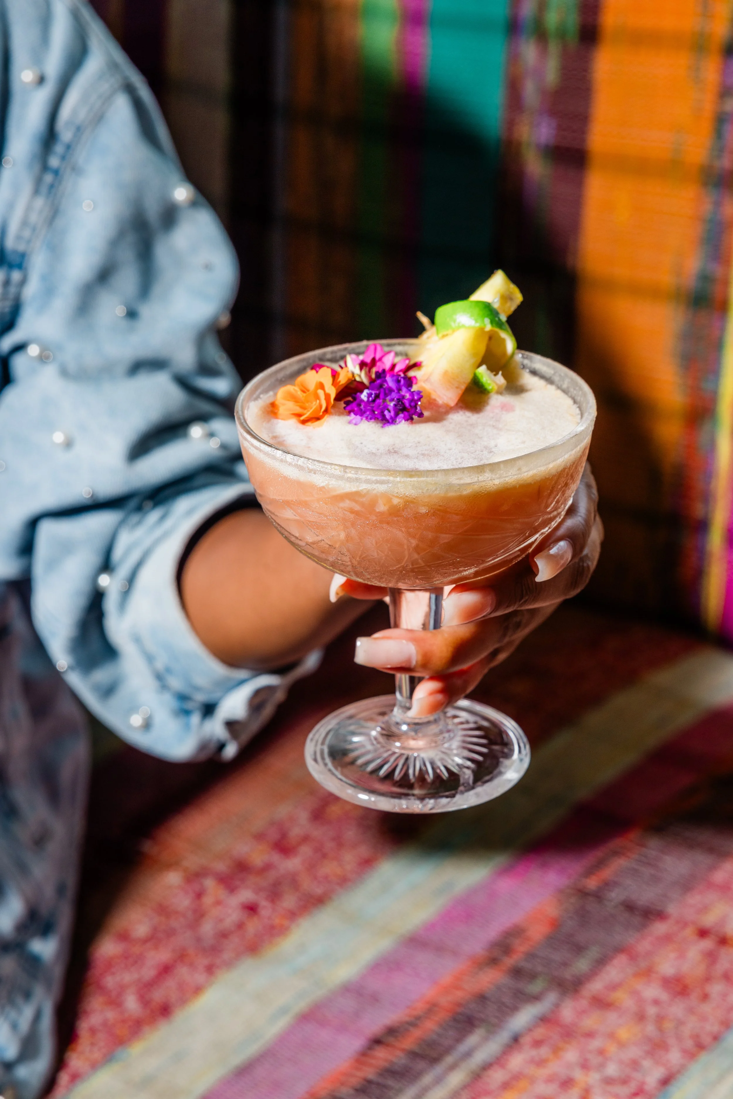 A person holding a pink cocktail garnished with fresh pineapple, lime, and edible flowers against a colorful woven fabric background.