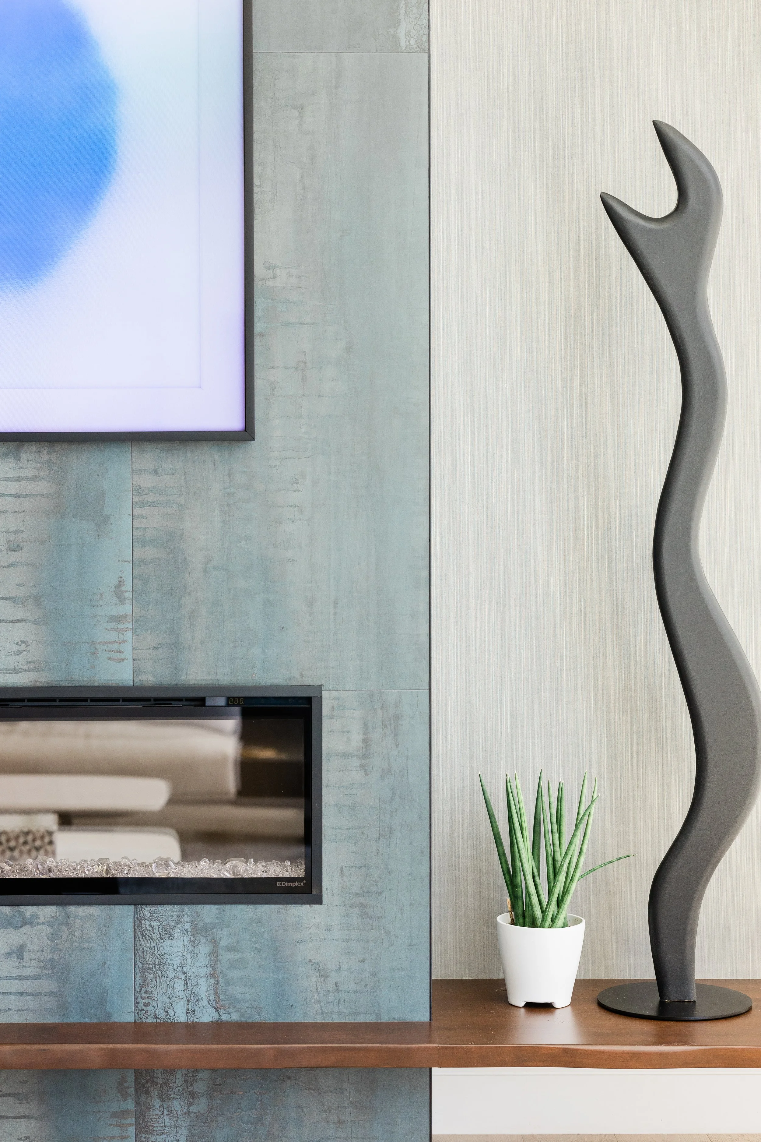 Interior of a modern living space with a flat-screen TV on a blue textured wall, a wooden shelf, a potted cactus plant, and a black abstract sculpture, with part of a fireplace below.