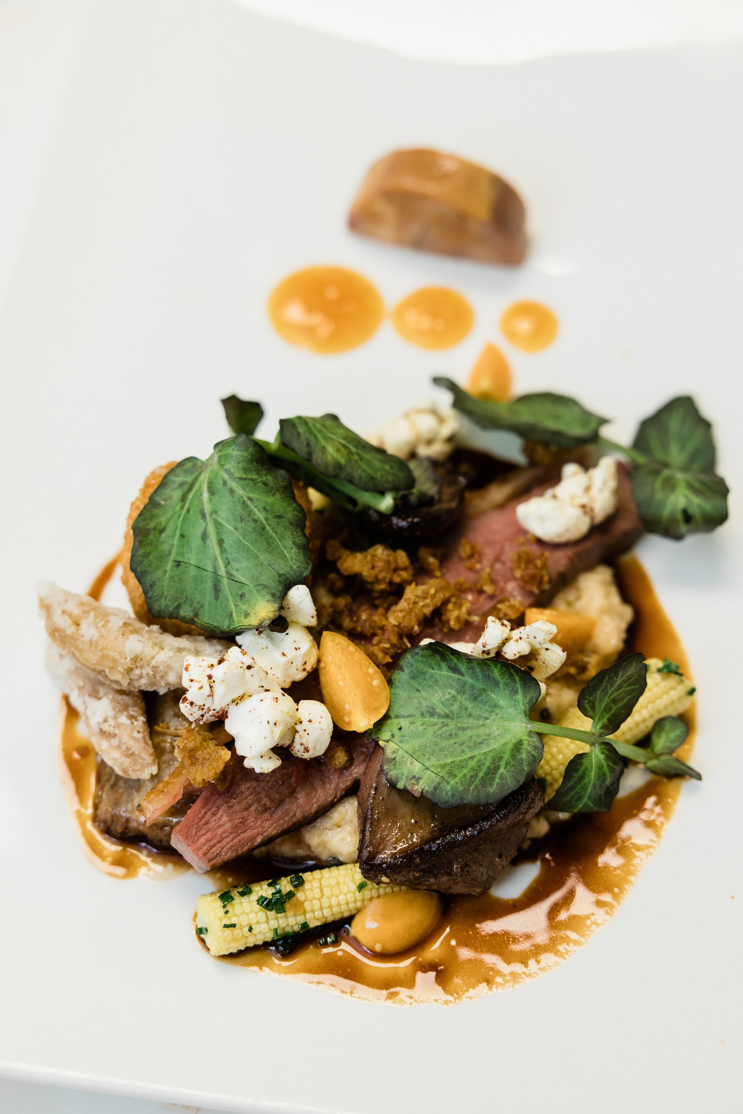 A gourmet dish featuring slices of medium-rare steak topped with fresh greens, popcorn, and a sauce, served with baby corn, peanuts, and a chocolate piece in the background.