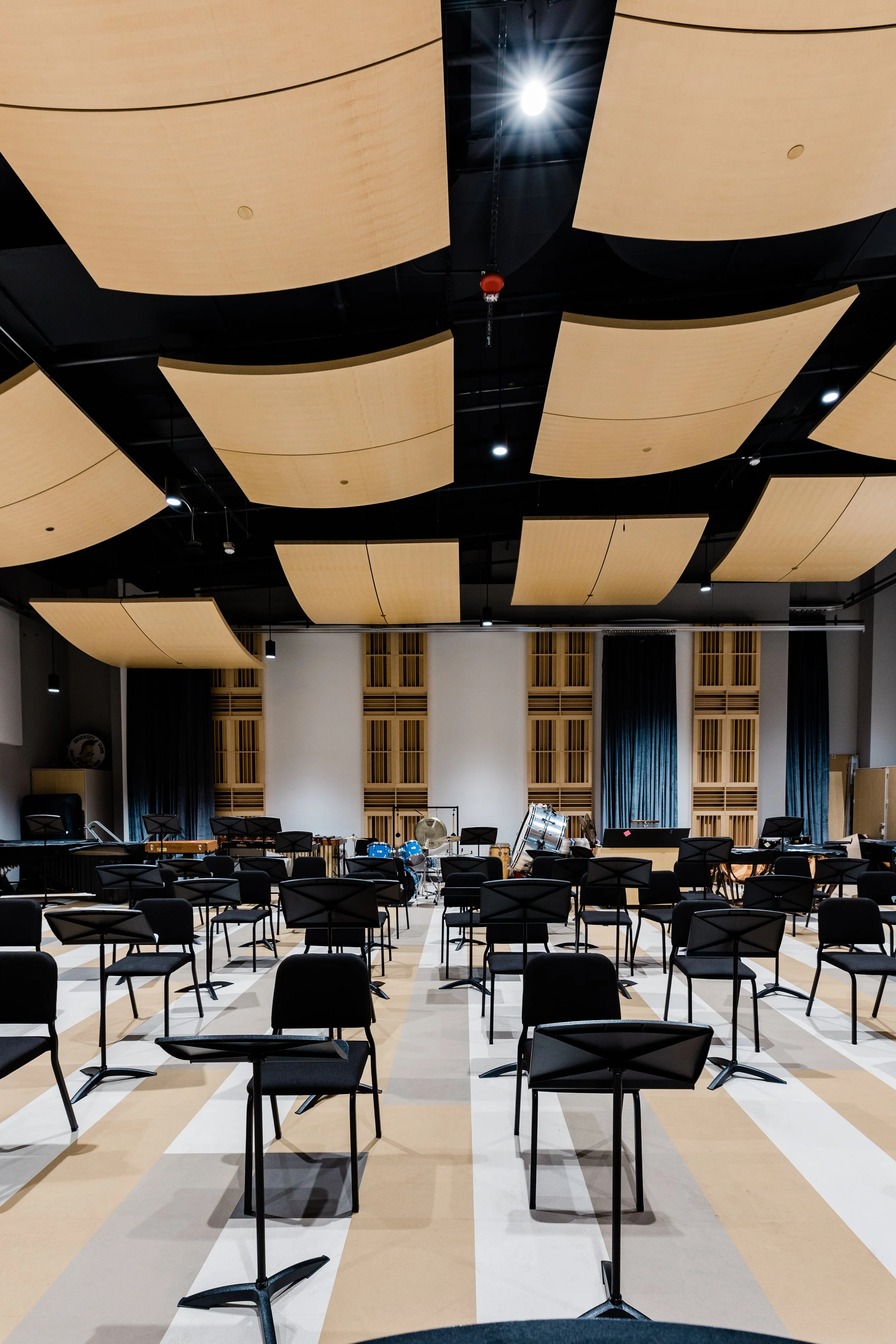 An empty concert hall or auditorium with black chairs arranged in rows facing a stage with musical instruments including drums, cymbals, and music stands. The ceiling has large, curved wooden panels and overhead lighting, and the back wall features s