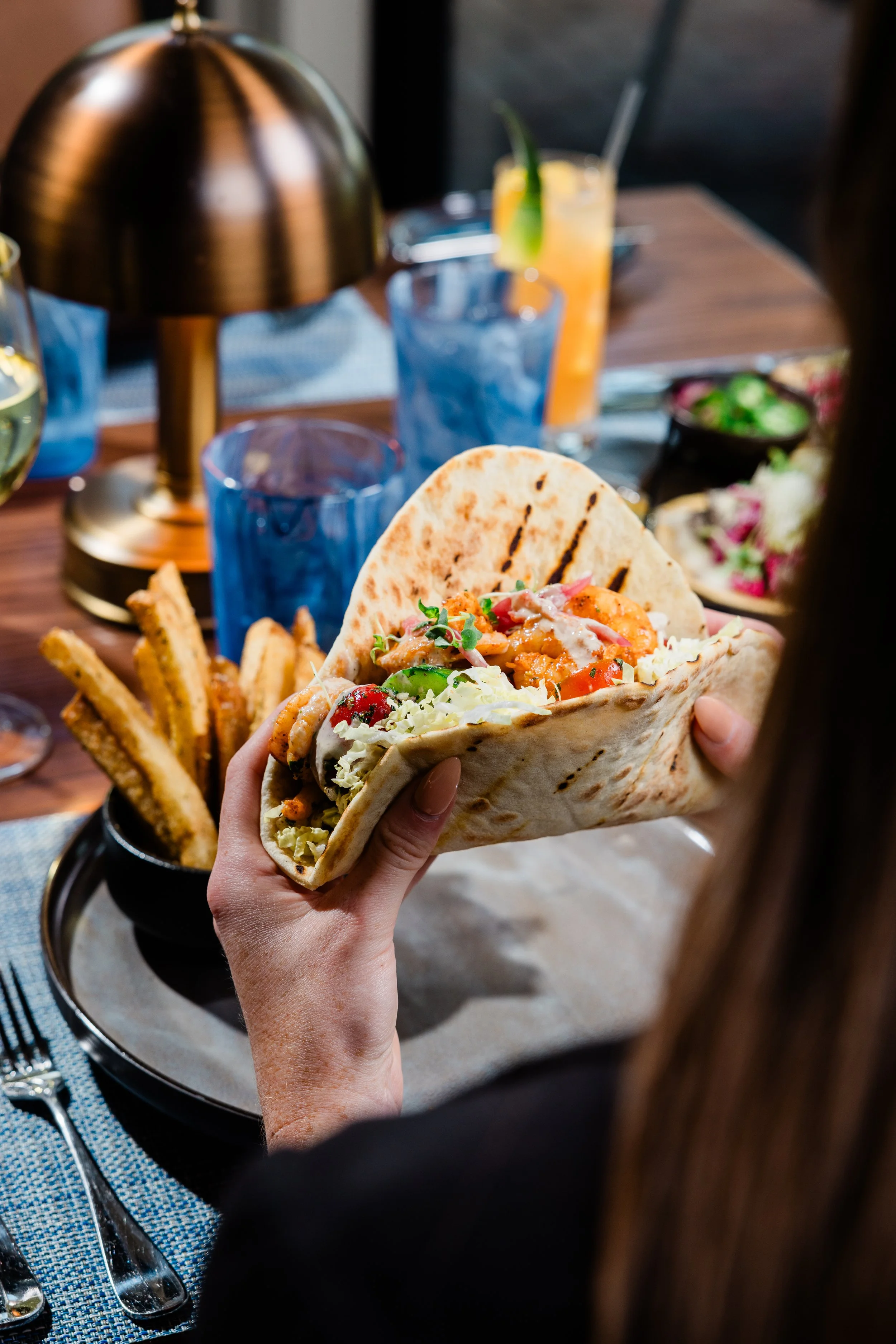 Person holding a taco with various fillings at a restaurant table, with drinks and fries nearby.