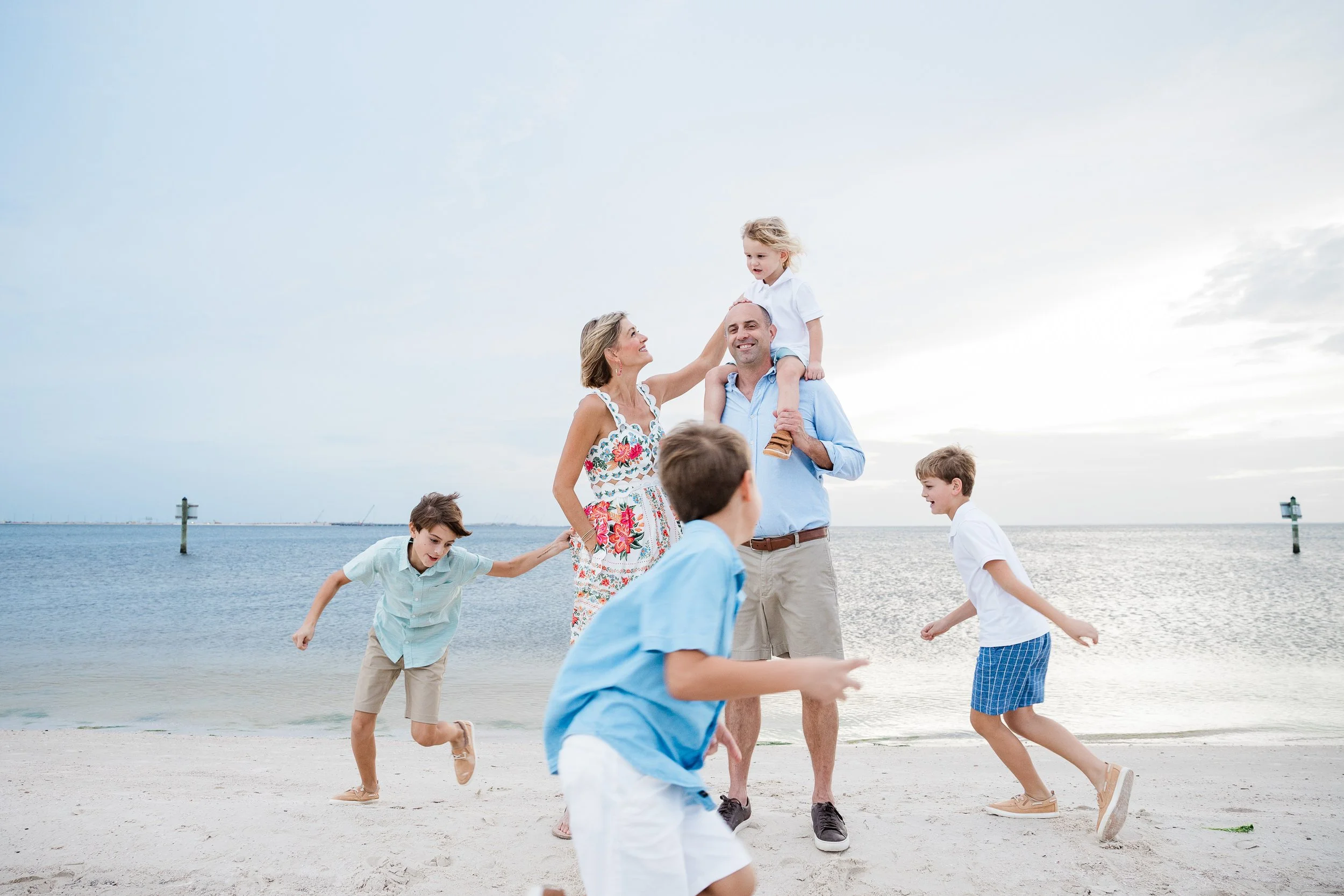 A family enjoying time together on a beach; a woman, man, and several children playing near the water under a cloudy sky.