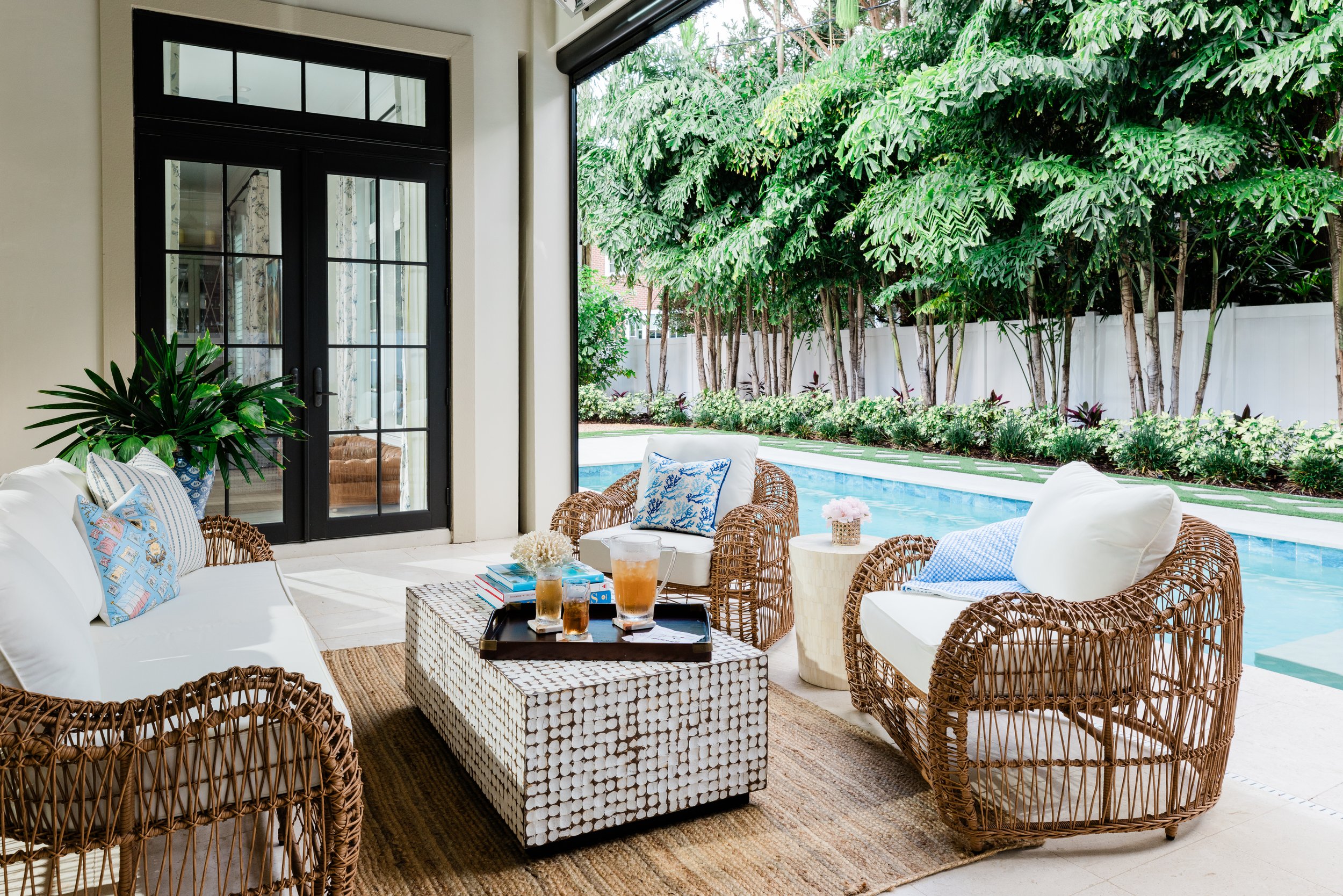 A covered outdoor patio with wicker furniture, cushions, and small tables, overlooking a lush garden and swimming pool. nterior design by J. Banks Design.