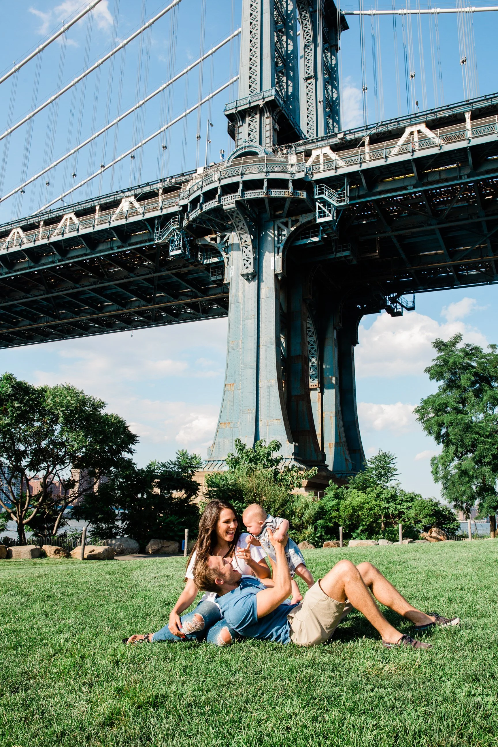 Family playing on the grass in a park beneath the Brooklyn Bridge on a sunny day with blue skies.