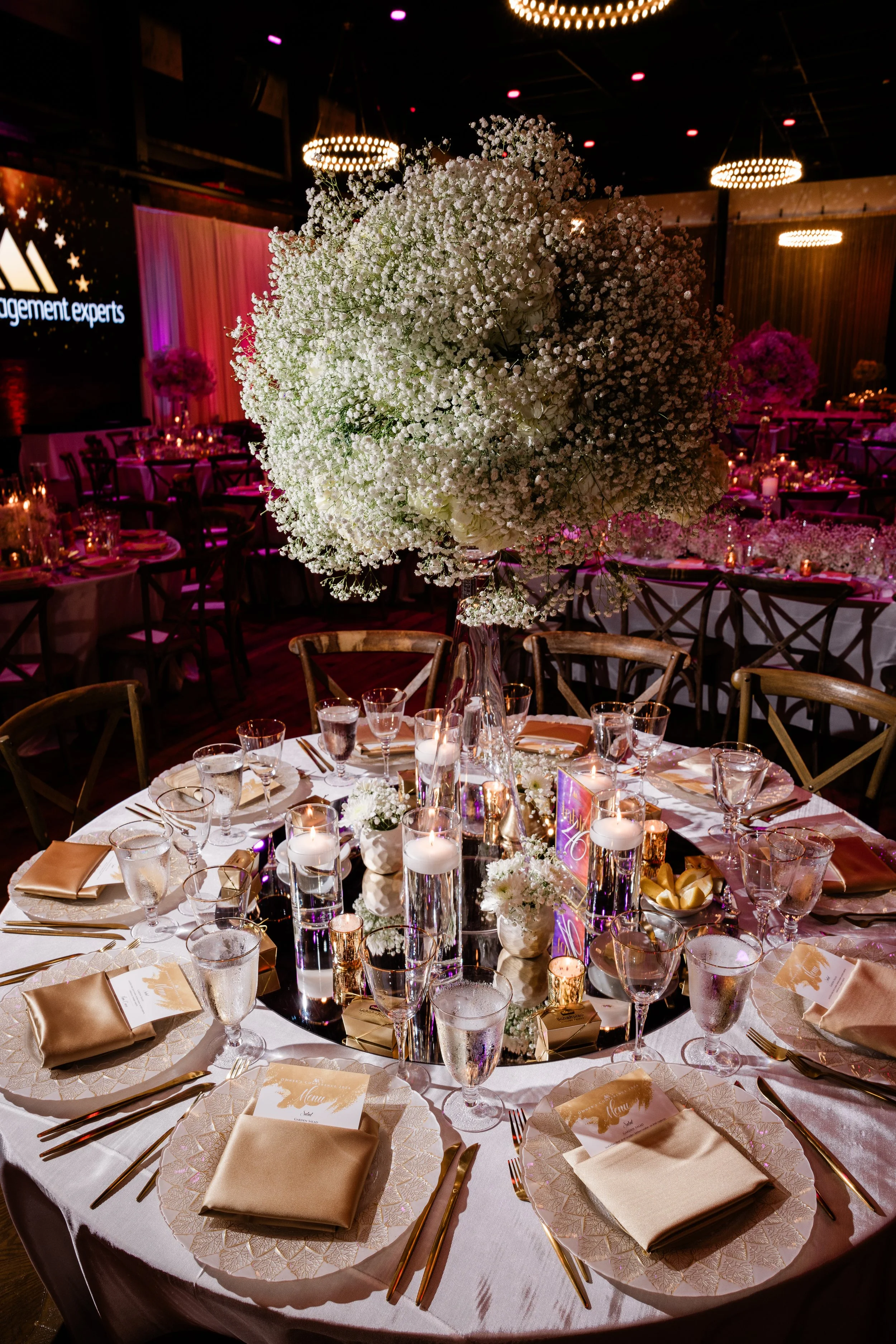 A round banquet table set for an event with a large floral centerpiece consisting of white flowers. The table is decorated with candles, glassware, gold-colored cutlery, and beige napkins on white plates. In the background, there are more tables, cha