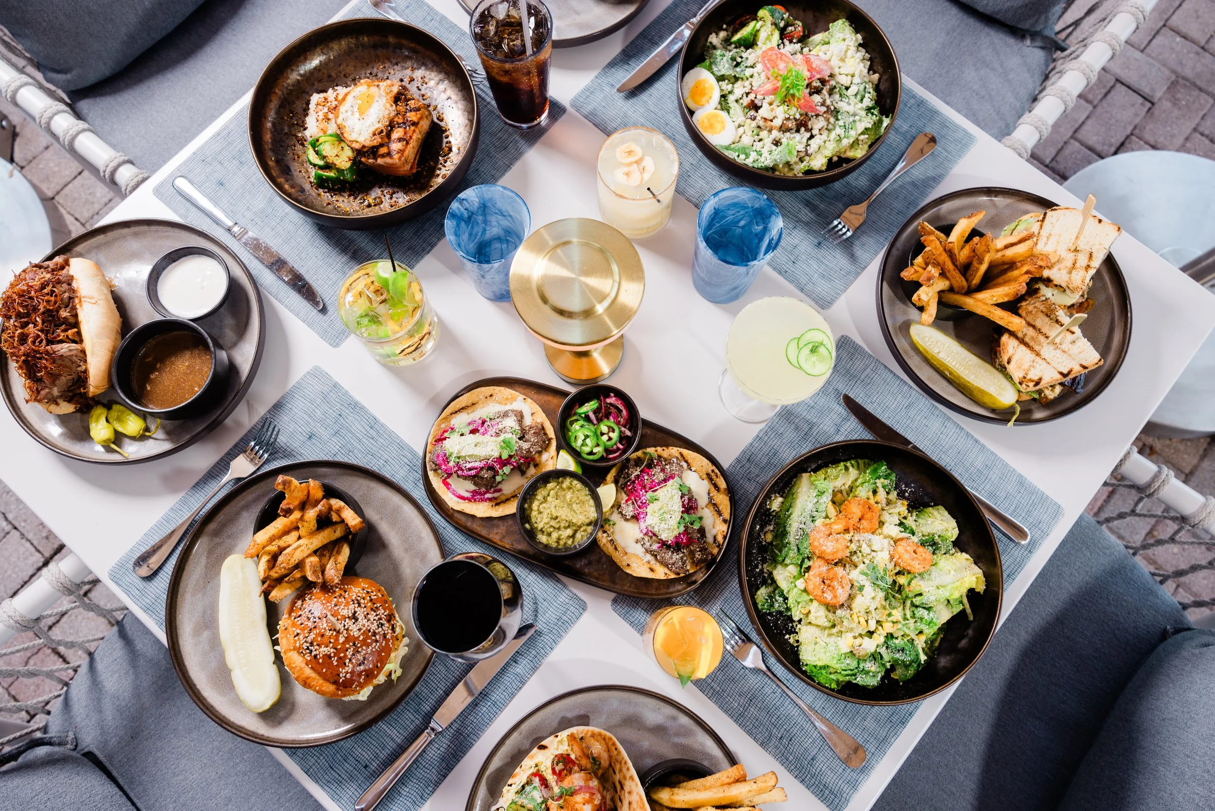 A top-down view of a table set with various plates of food including salads, sandwiches, tacos, a burger, and drinks, on a white table with blue placemats.