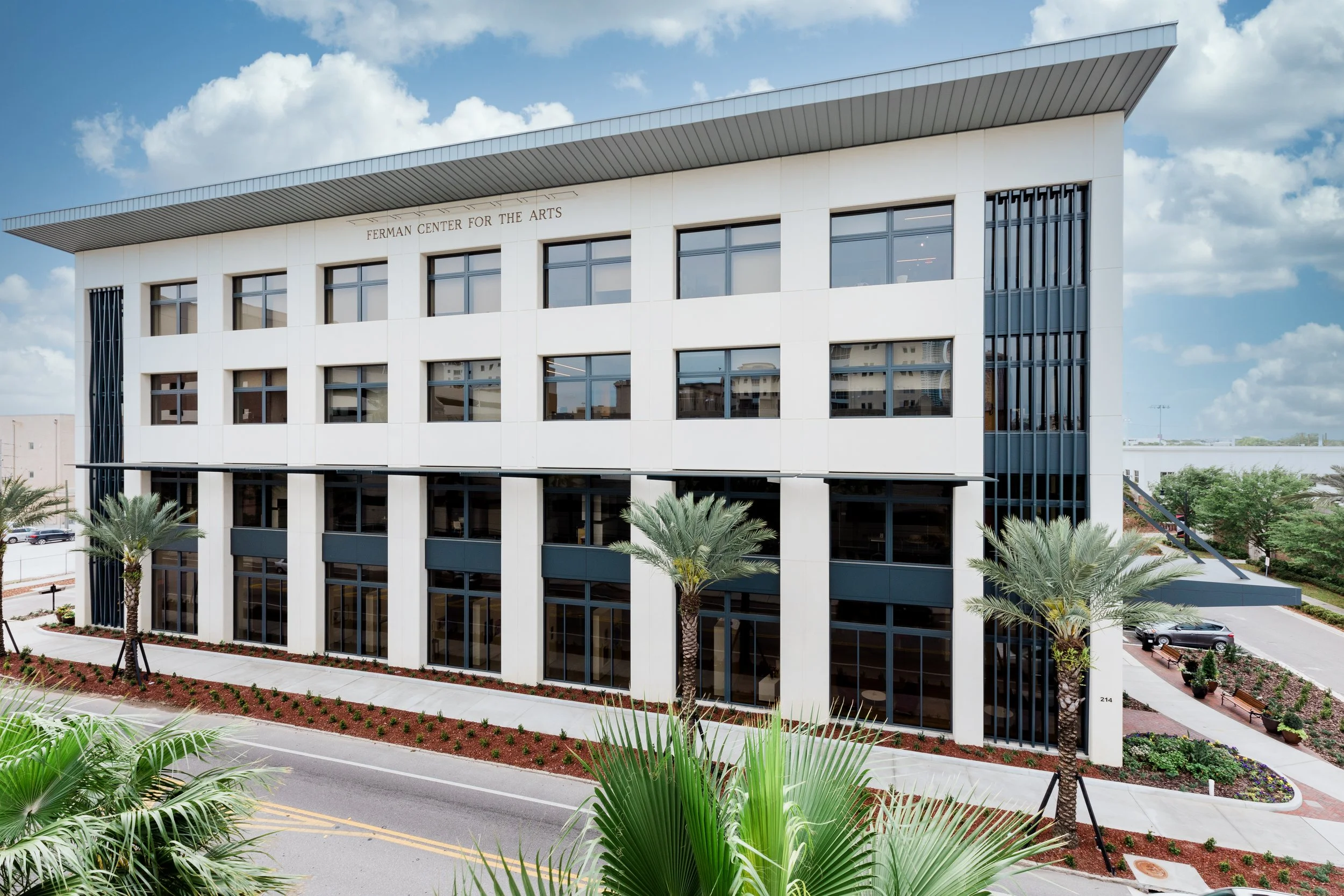 Modern white building with multiple large windows, labeled 'Ferman Center for the Arts,' surrounded by landscaping including palm trees and flower beds, under a partly cloudy sky.