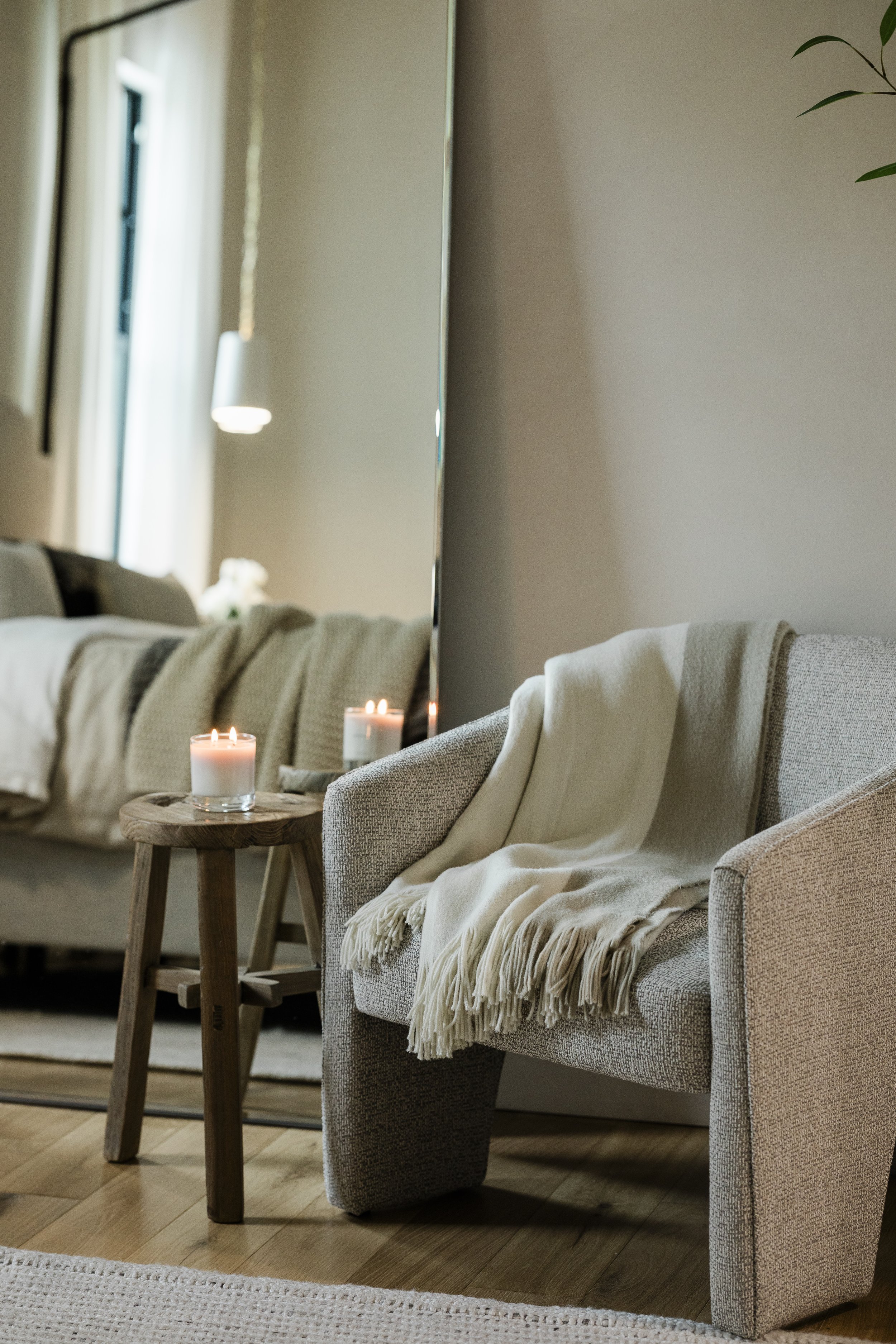 Living room with beige armchair, white throw blanket, small wooden side table with candles, beige sofa with beige and brown throw blanket, large mirror on wall reflects window with curtains, candles on table and sofa, wooden flooring.