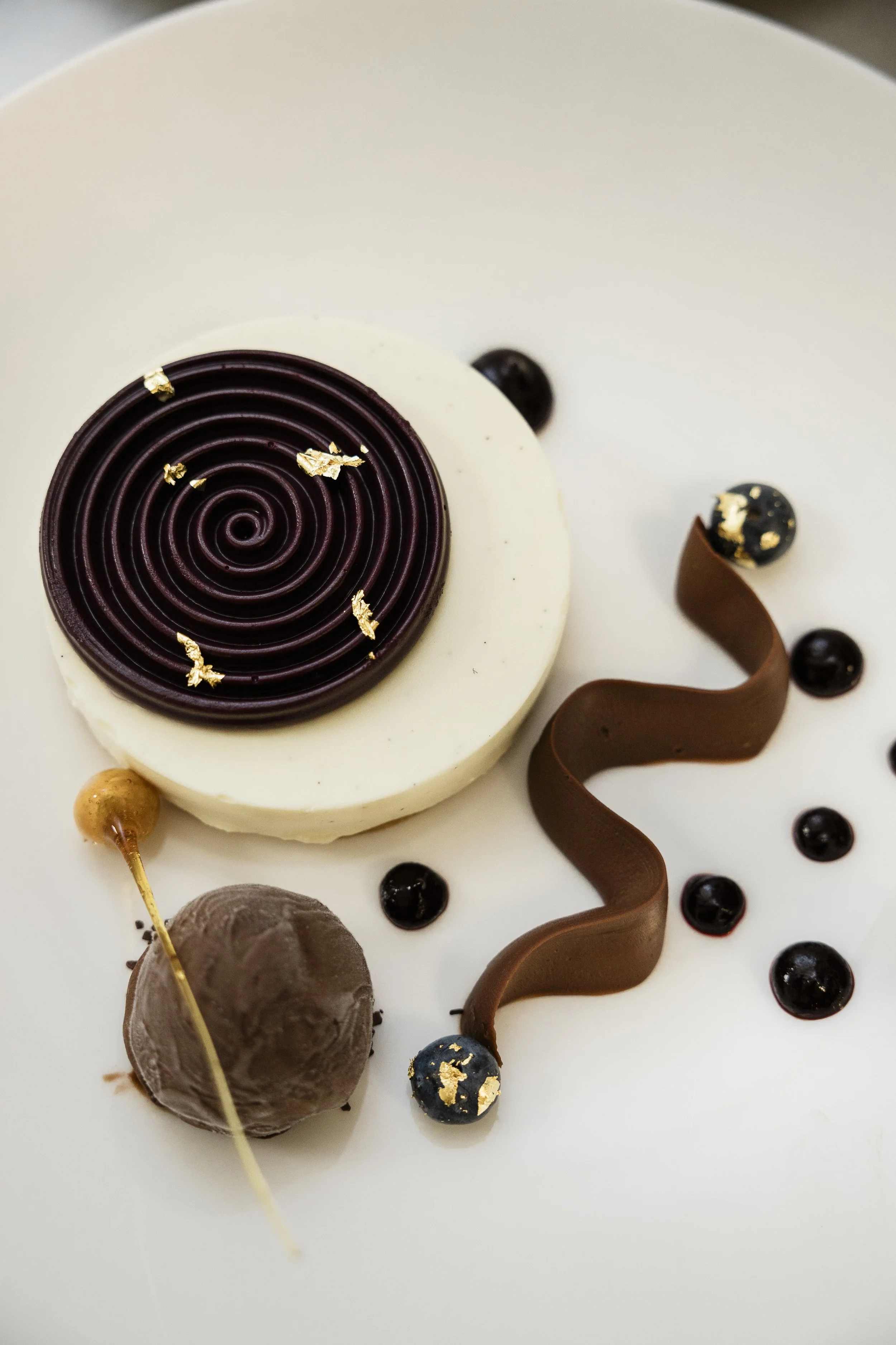 Close-up of a decorated birthday cake with a chocolate spiral design, gold leaf accents, chocolate swirls, black and gold spherical decorations, and a small chocolate ball with a skewer.