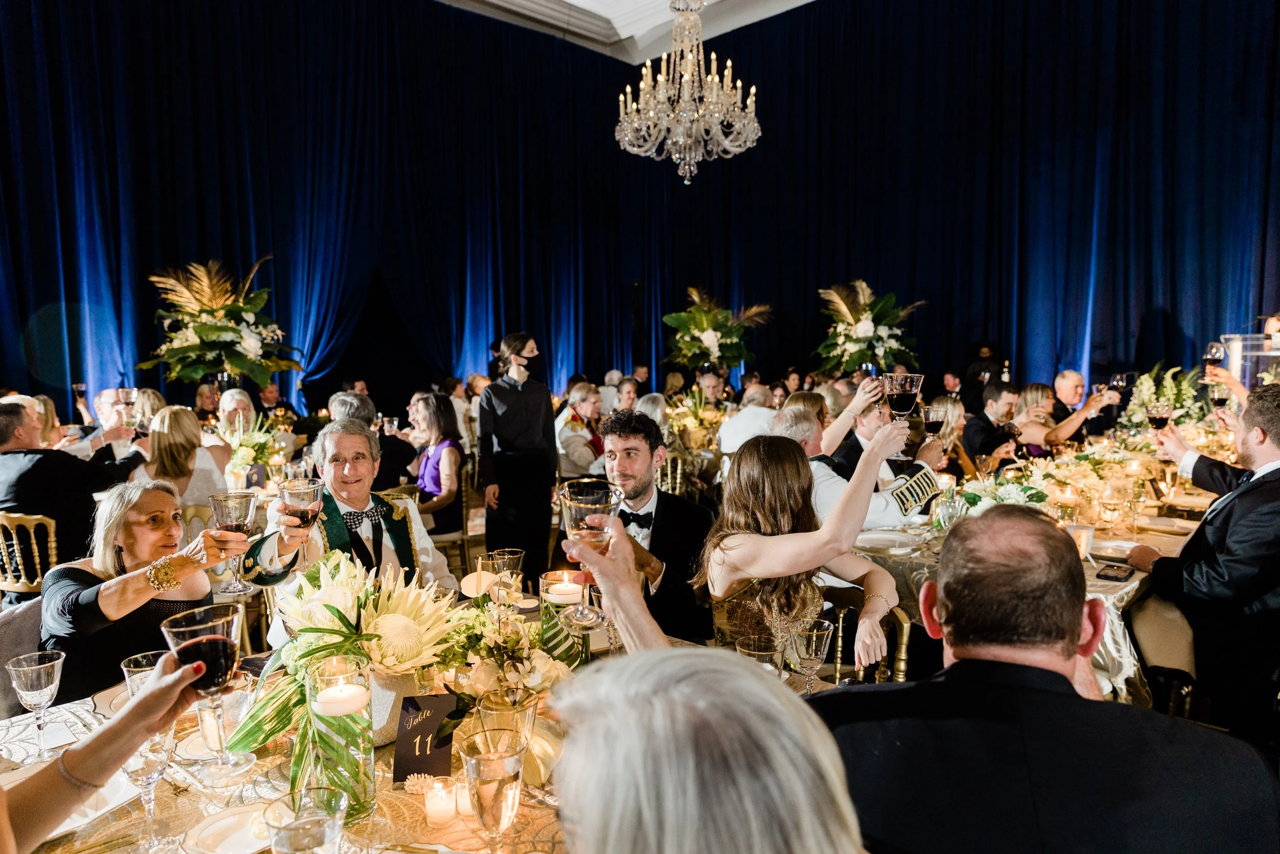 Guests at a formal dinner celebration raise glasses for a toast in an elegant ballroom with a large chandelier, blue curtains, and floral centerpieces.