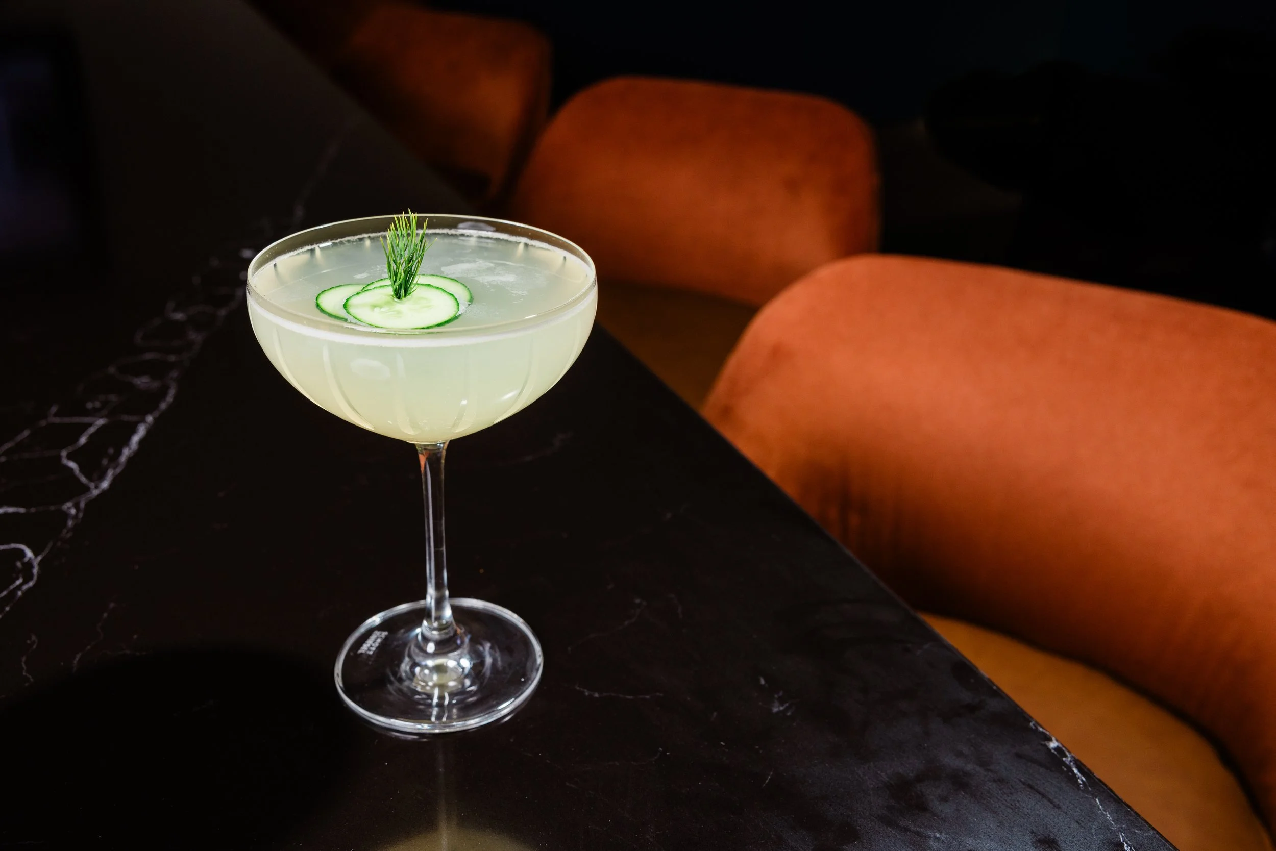 A cocktail glass filled with a light green beverage, garnished with thin cucumber slices and a sprig of dill on top, placed on a black marble surface with orange chairs in the background.
