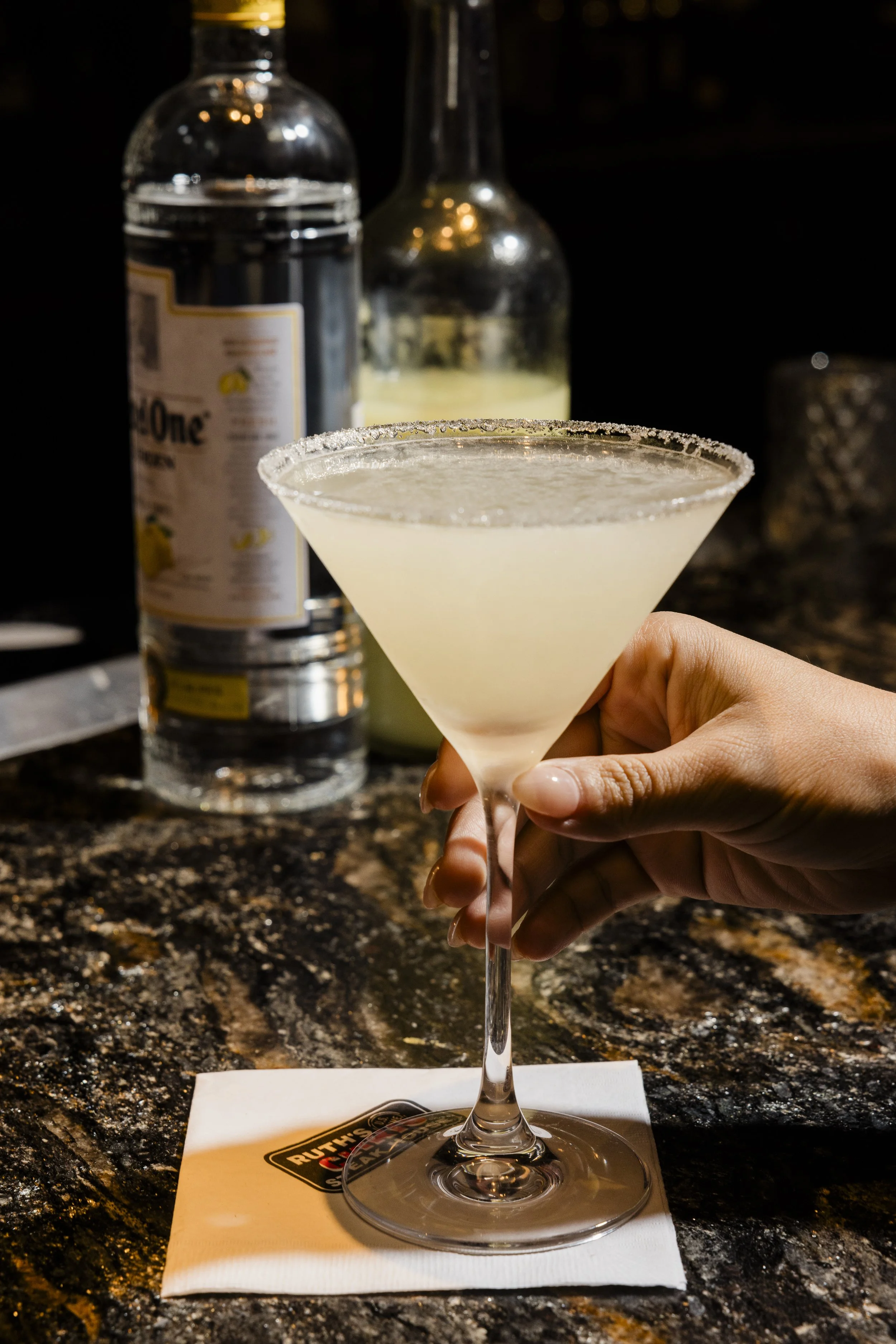 A bartender's hand holding a lemon drop martini glass with a sugared rim, on a napkin on a dark, granite bar counter, with a bottle of tonic water and a bottle of alcohol in the background.