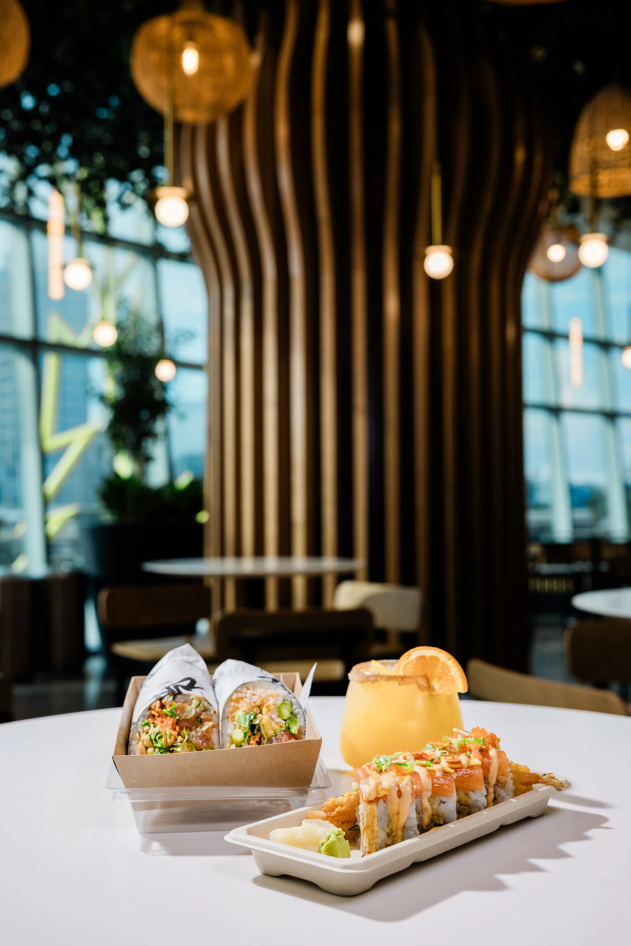 A table featuring two sushi rolls in a paper tray, a small dessert topped with orange slices and sauce, and a small container of wasabi and ginger, set against a blurred background of a modern, well-lit restaurant interior with large windows and ambi