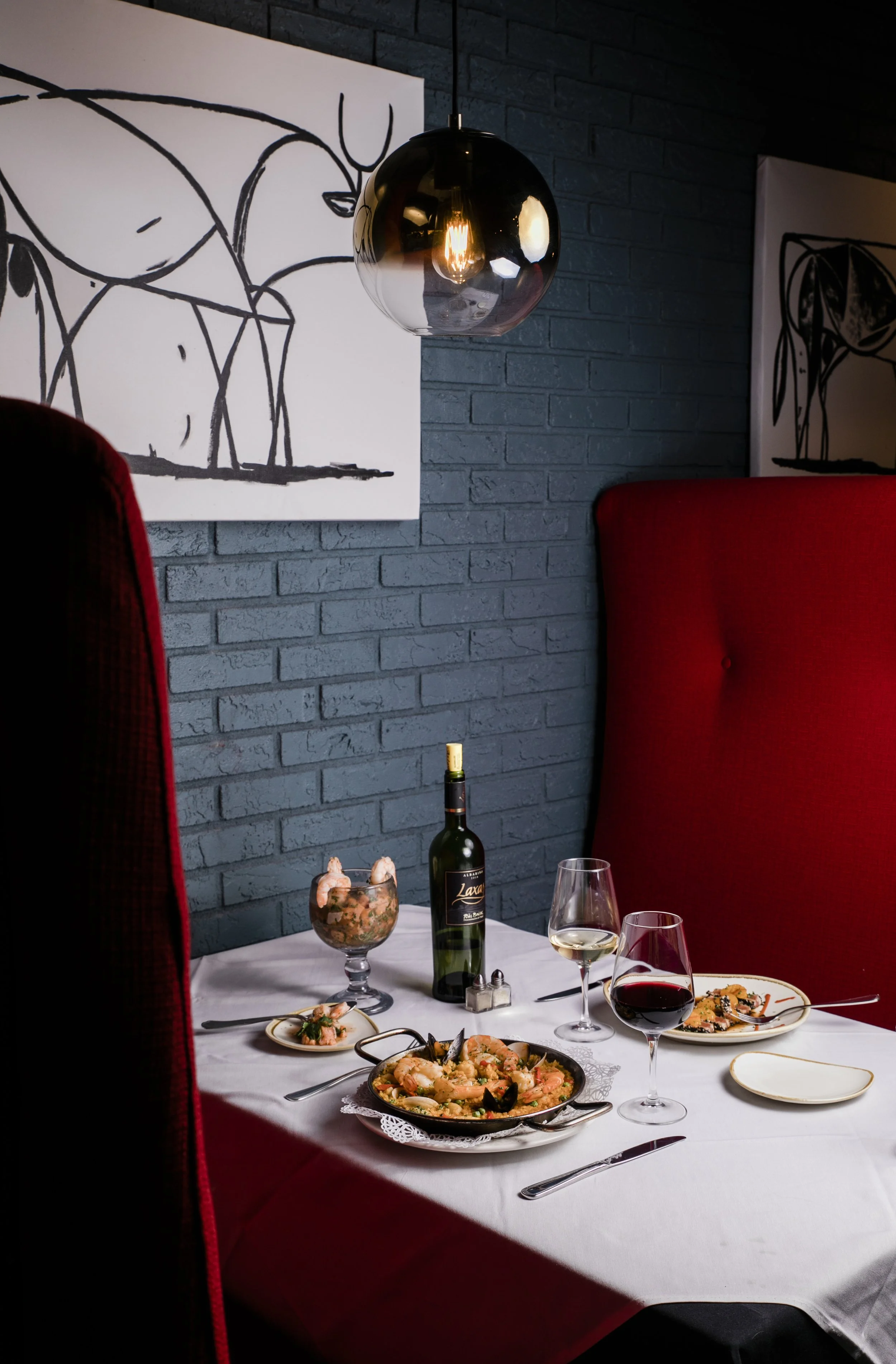 Dinner set for two on a white tablecloth next to grey brick wall with modern sketch of cow. Red banquette. Vizcaya Restaurant.
