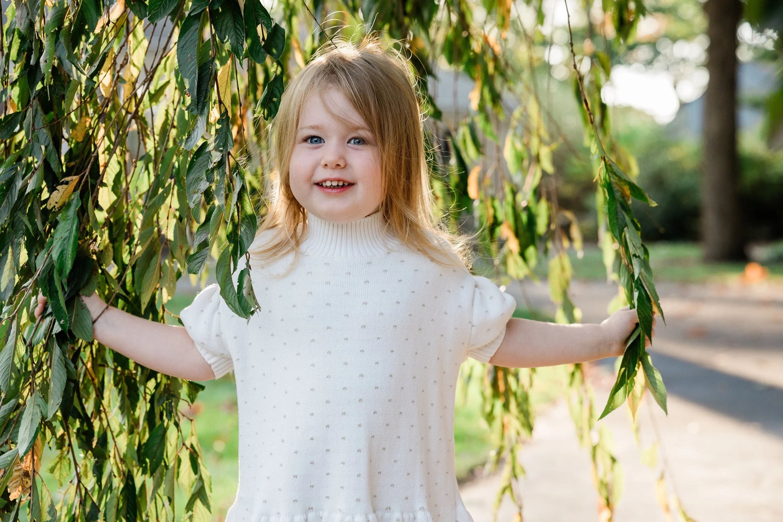 A young girl with blonde hair and blue eyes standing outdoors during daytime, holding green leafy branches on each side, with a blurred background of trees and a pathway.