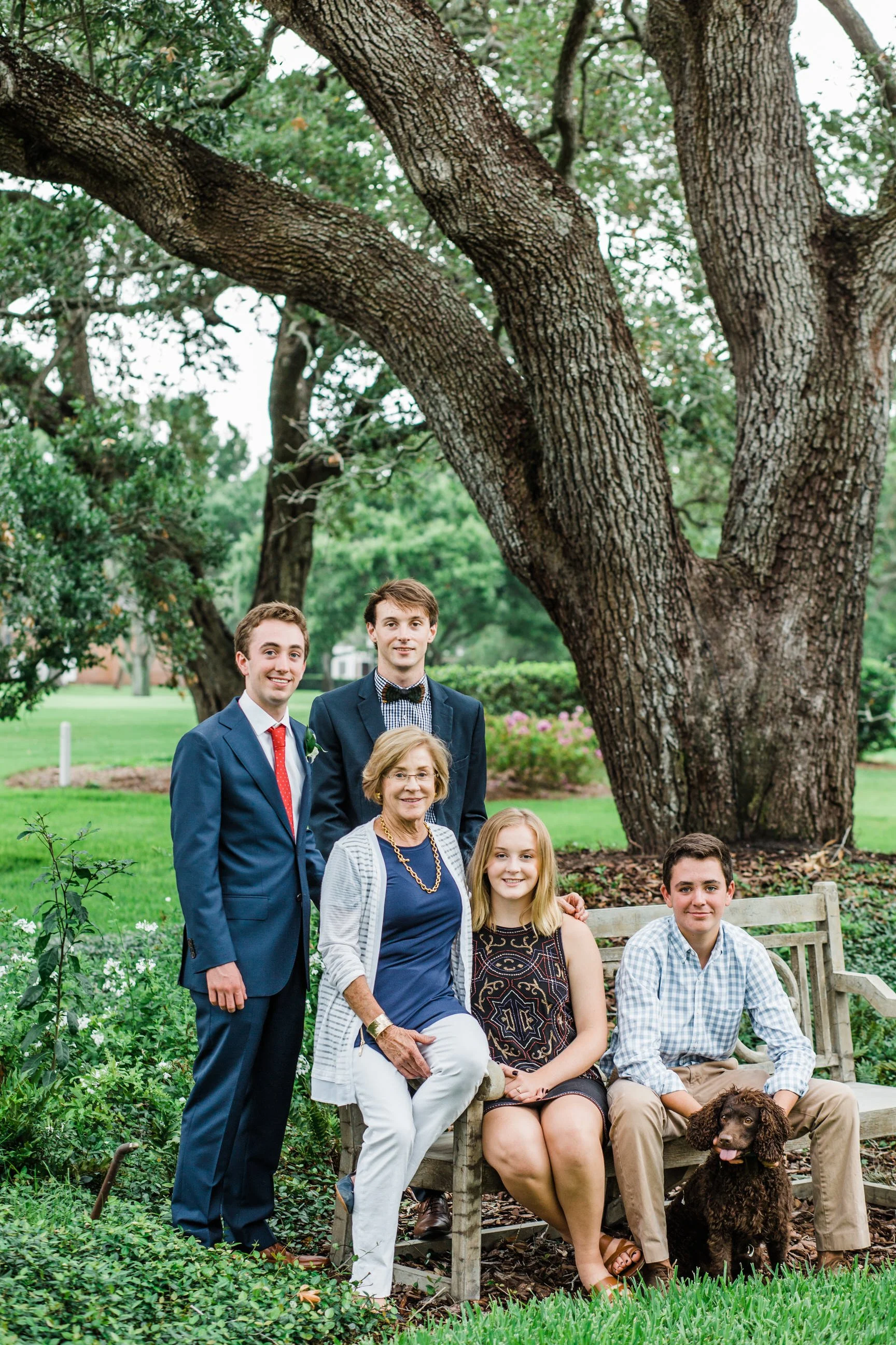 A group of seven people, including children and adults, posing outdoors in a garden with greenery and large trees, along with a brown dog, some sitting on a bench and others standing.