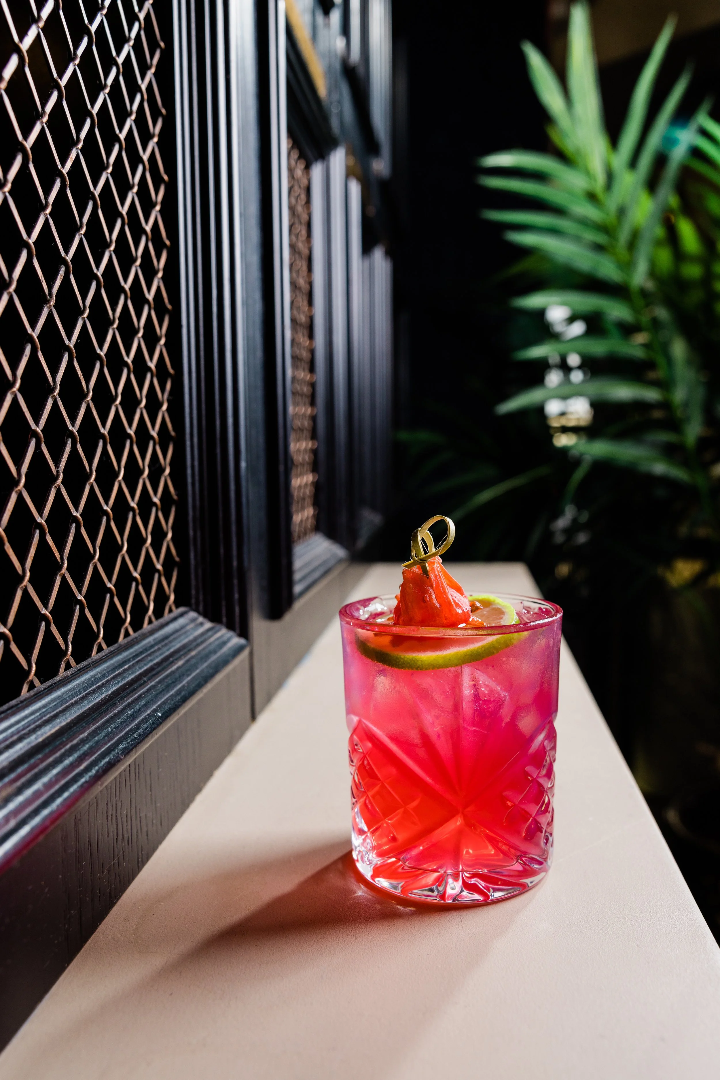 A pink cocktail in a textured glass with slices of lime and a dollop of red fruit or sauce on top, placed on a white surface near a dark wall with a black metal grid and green plants in the background.