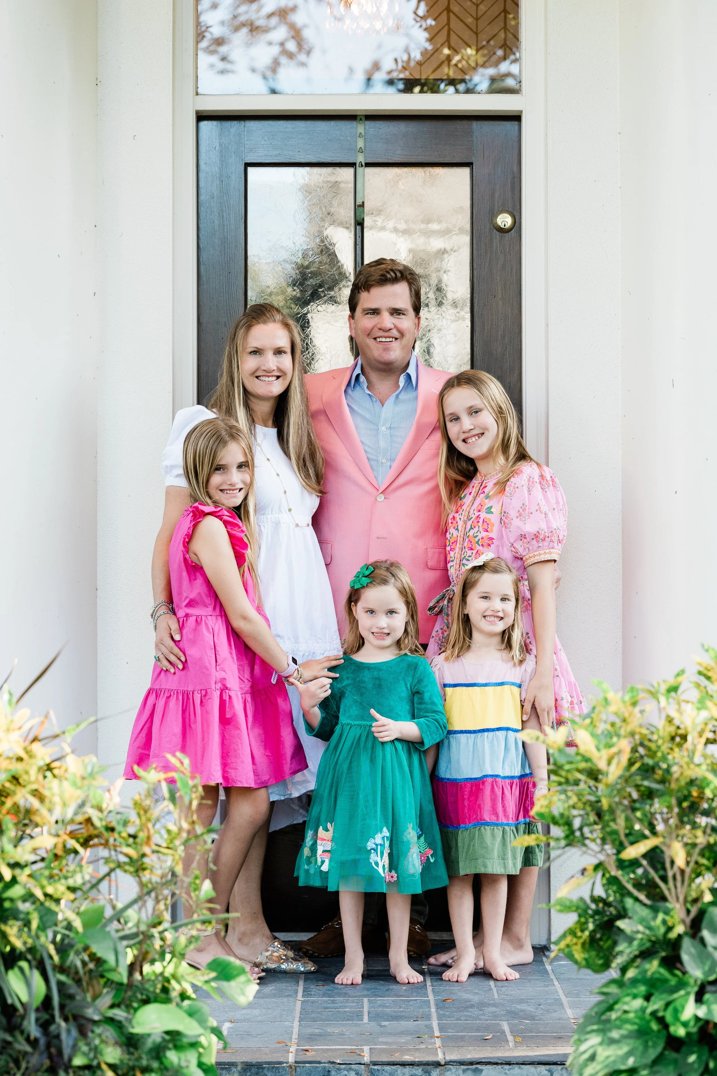 A family of six standing on a porch in front of a dark door with a glass panel. The family includes a man in a pink blazer, a woman in a white dress, and four young girls in colorful dresses. The family is smiling for the photo, with some of the girl