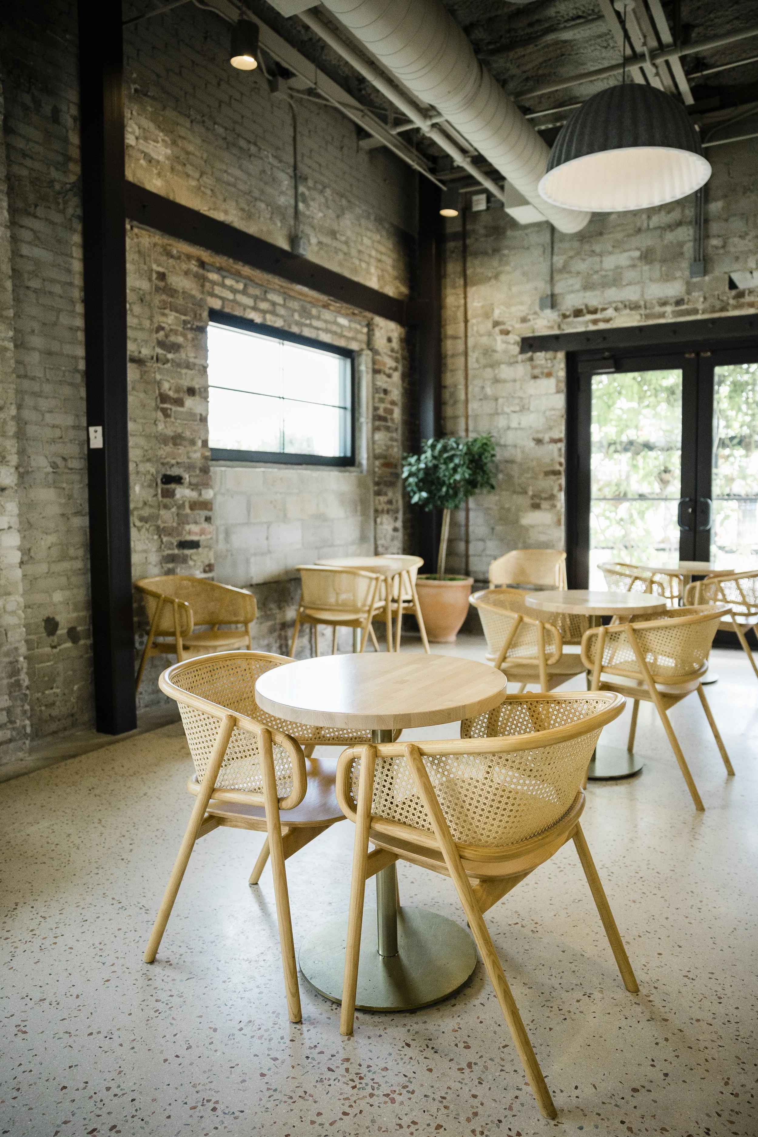 Interior of a modern cafe with exposed brick walls, circular wooden tables, and woven wooden chairs. Large windows and a glass door let in natural light, and a potted plant is visible in the corner. Industrial-style ceiling with visible ductwork and 