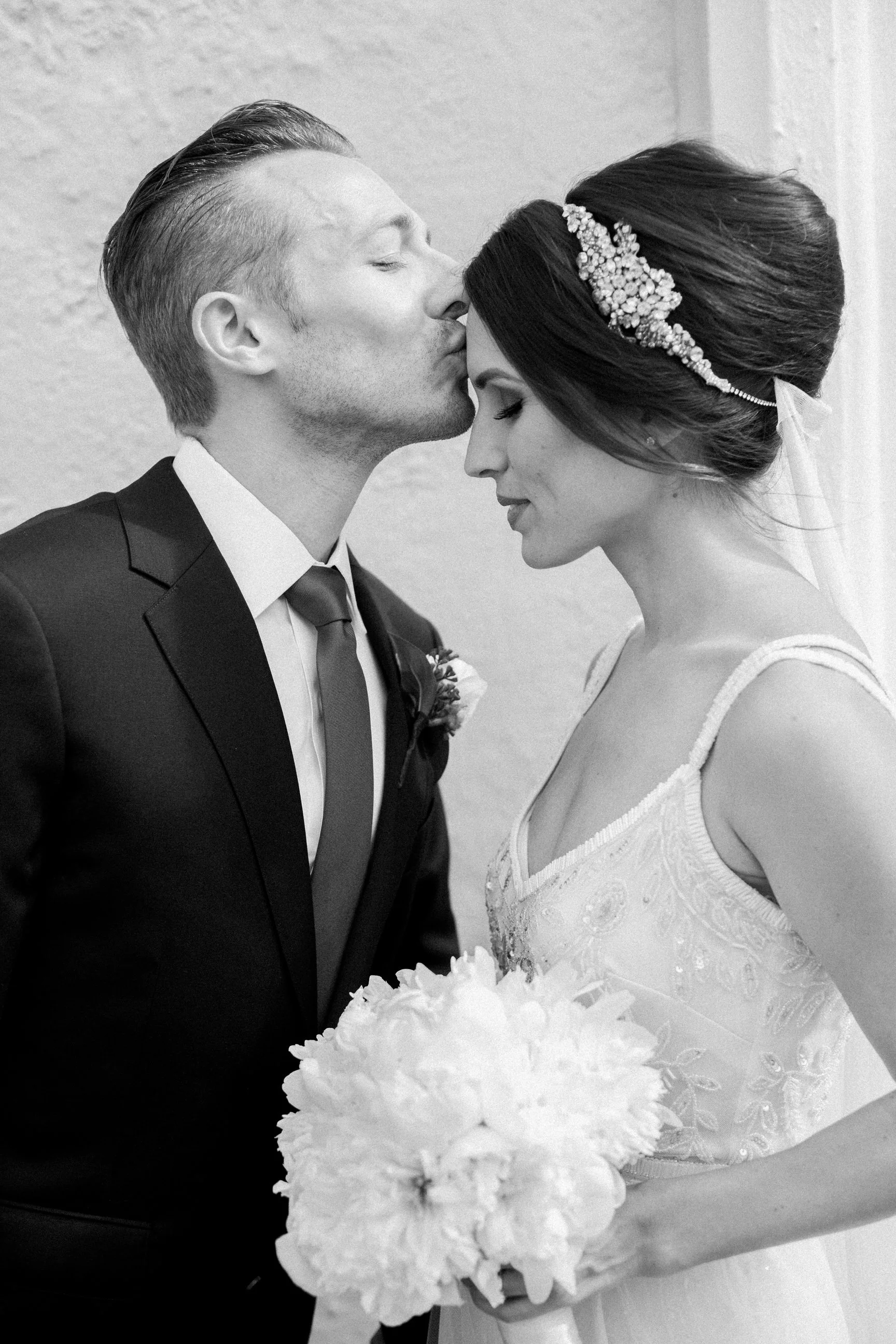 A black and white photo of a bride and groom, with the groom kissing the bride's forehead. The bride has her eyes closed and is holding a bouquet of flowers. The groom is dressed in a suit and tie, and the bride is wearing a wedding dress with a flor
