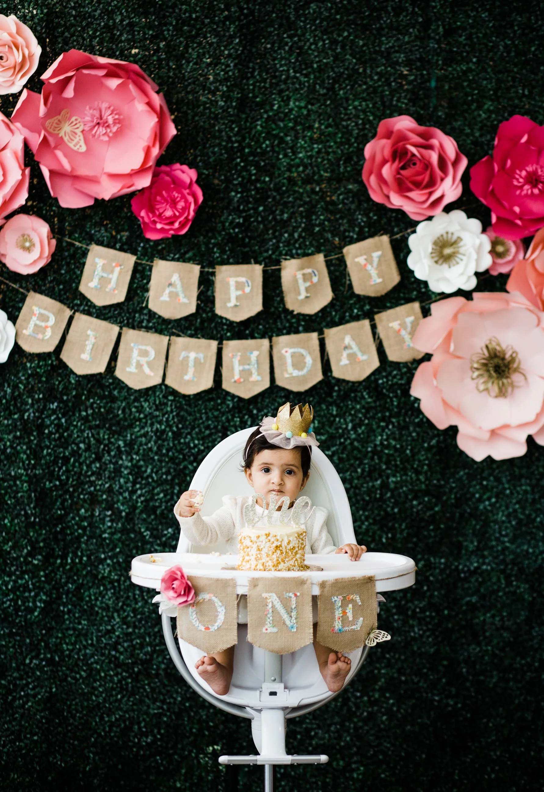 A young child sitting in a high chair with a birthday cake in front of her, wearing a birthday crown and a white sweater, with a birthday banner and large pink and white paper flowers on a dark green textured background
