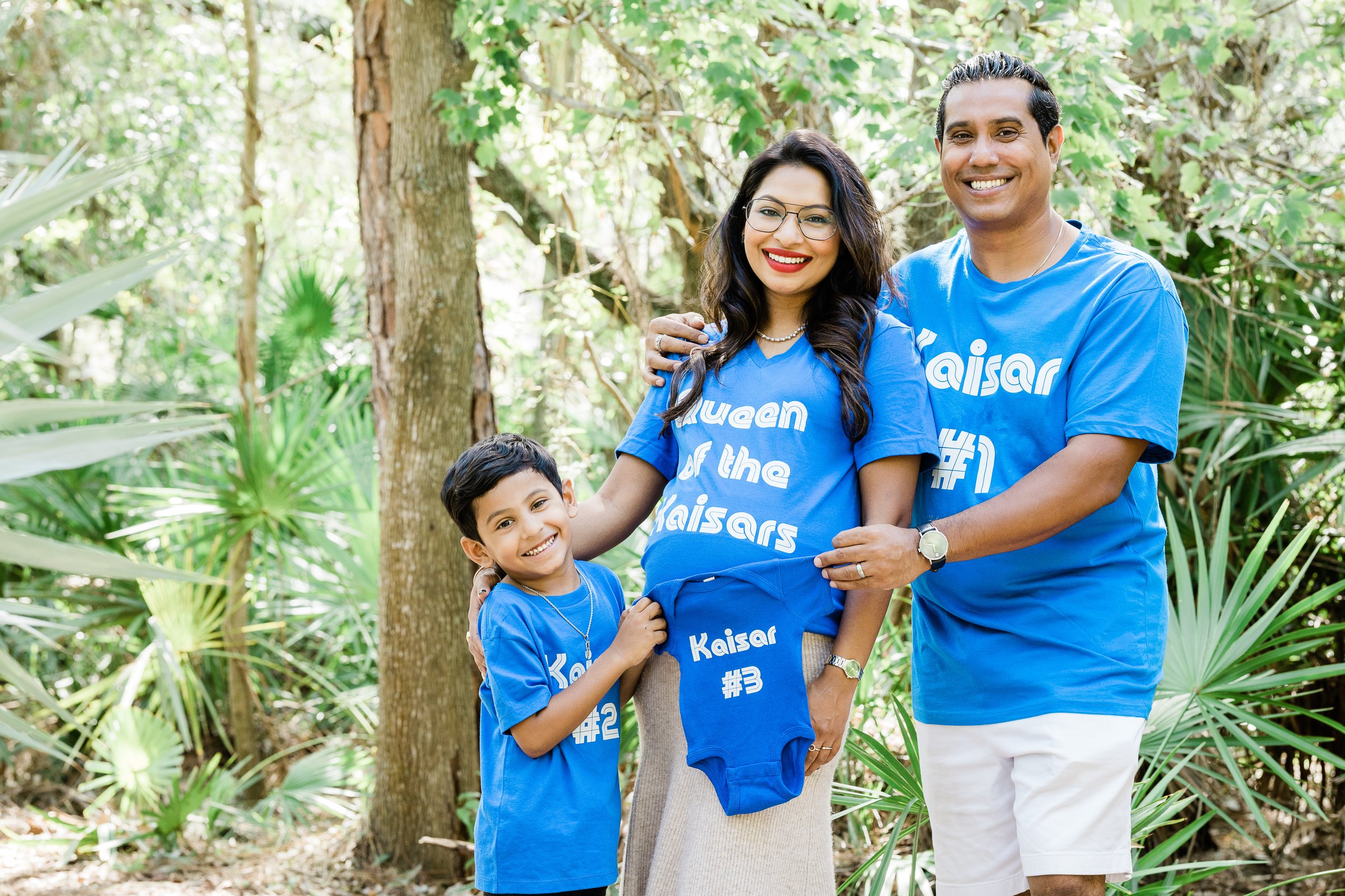 A smiling family of four standing outdoors among trees and greenery, wearing matching blue t-shirts with slogans and numbers, posing for a photo.