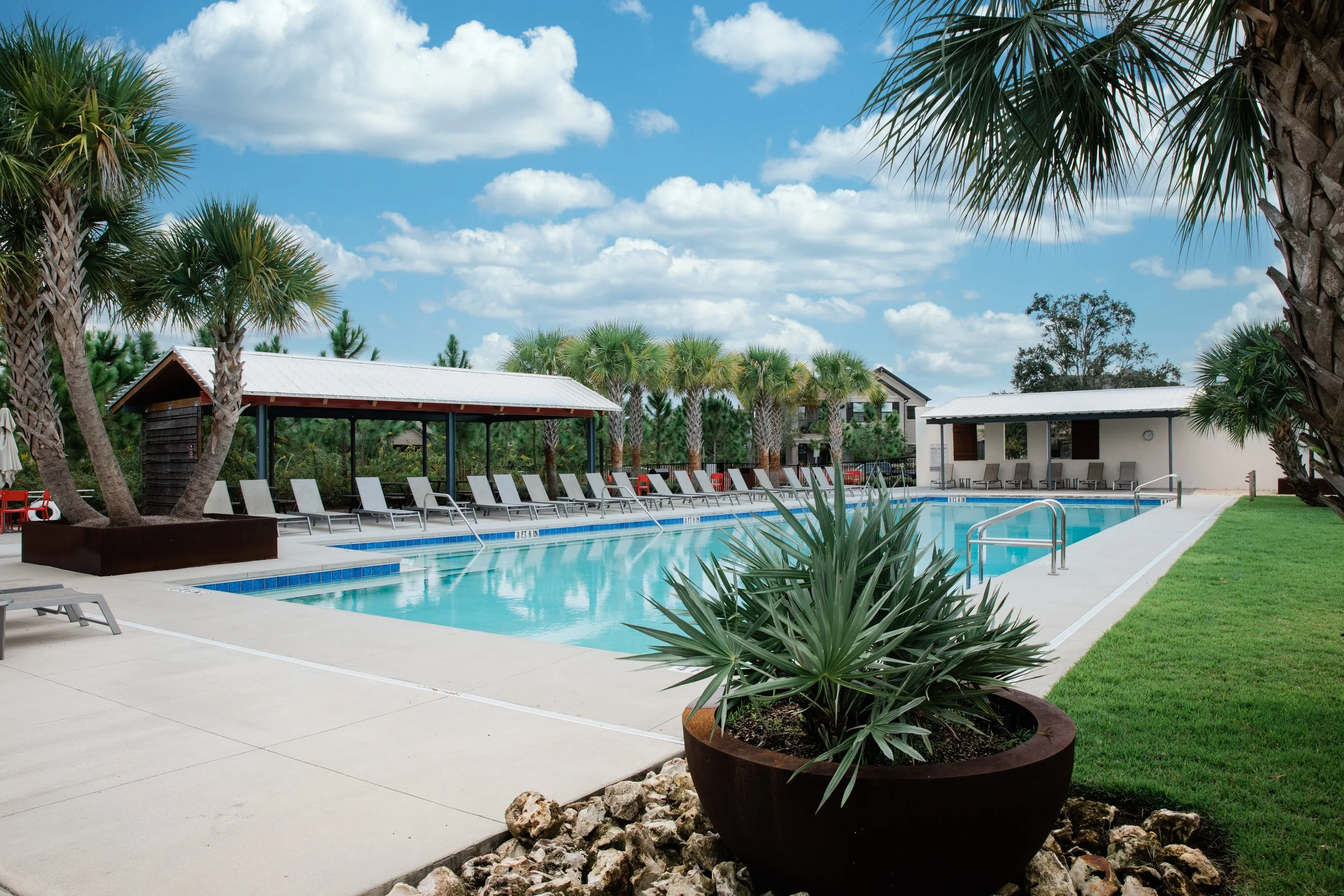 Starkey Ranch pool detail. Potted plant in foreground, outdoor pool with accessory buildings in background. Blue sky with fluffy white clouds. Designed by Mike Ruble.