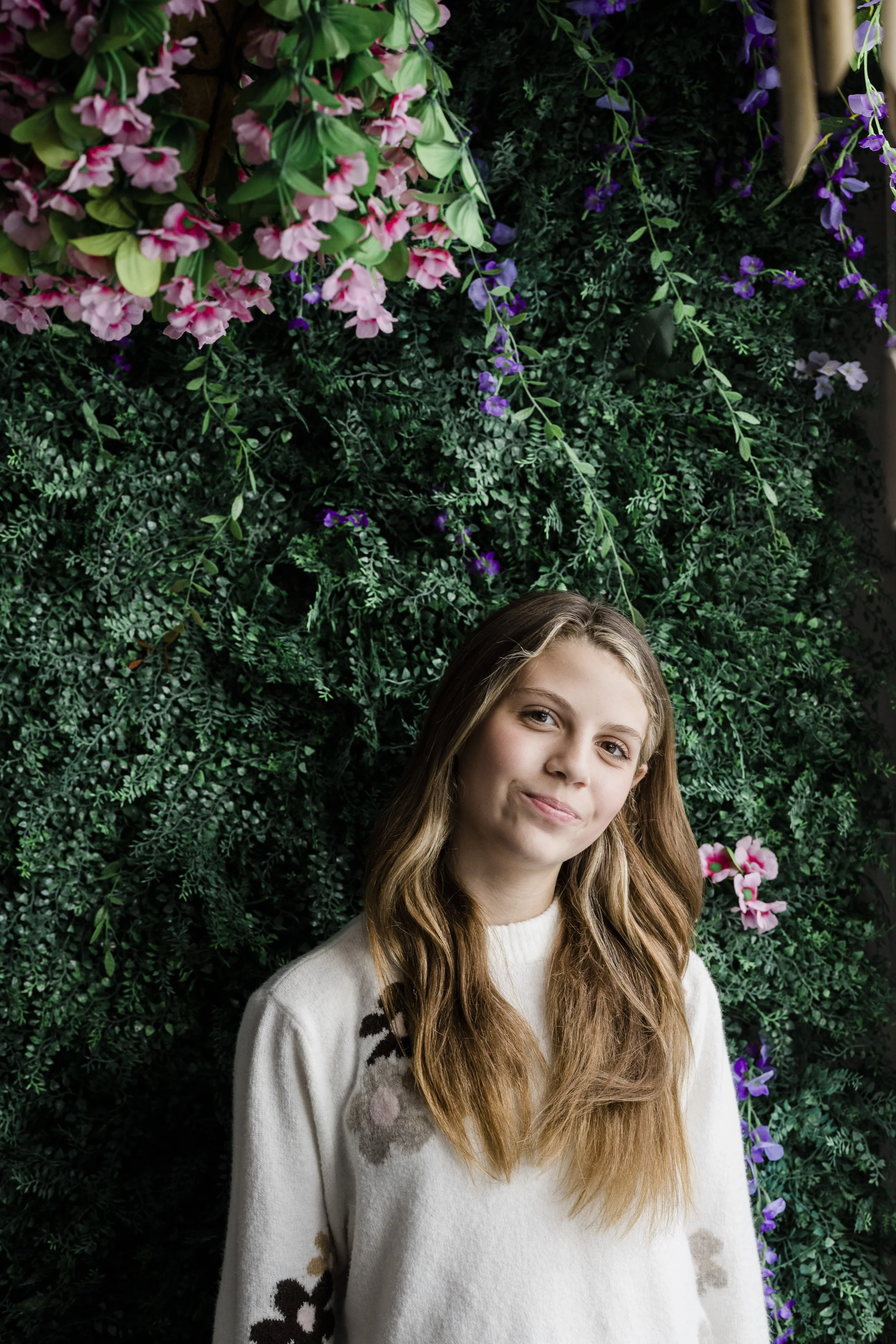 A young girl with long, wavy brown hair and light skin is smiling softly at the camera. She is wearing a white sweater with floral patterns on the sleeves. Behind her is a lush green vine wall with pink and purple flowers.