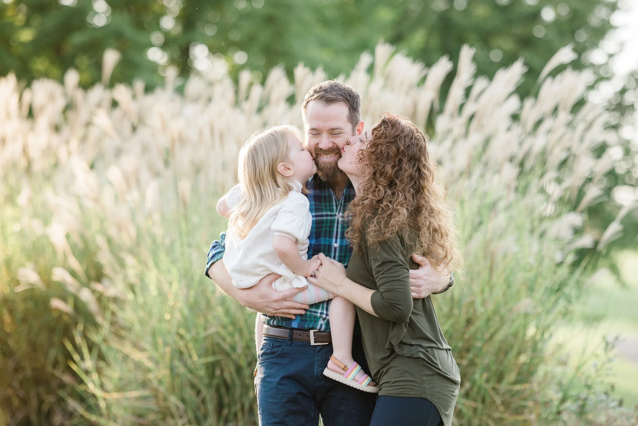 A happy family moment outdoors showing a man, woman, and young girl sharing kisses on the cheeks.