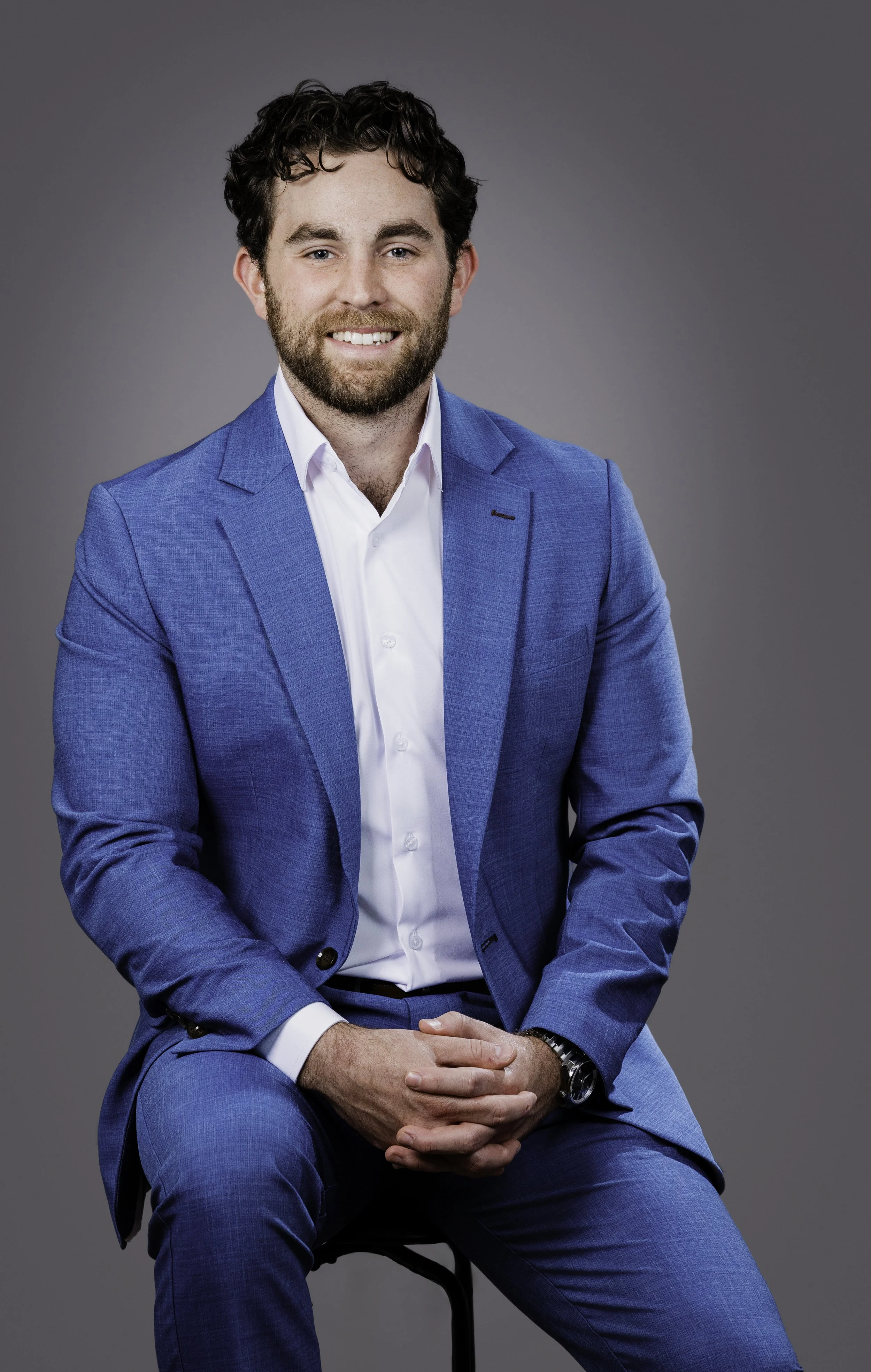 A man with dark curly hair, a beard, and light skin, smiling, wearing a bright blue suit with a white shirt, sitting on a stool against a plain grey background.