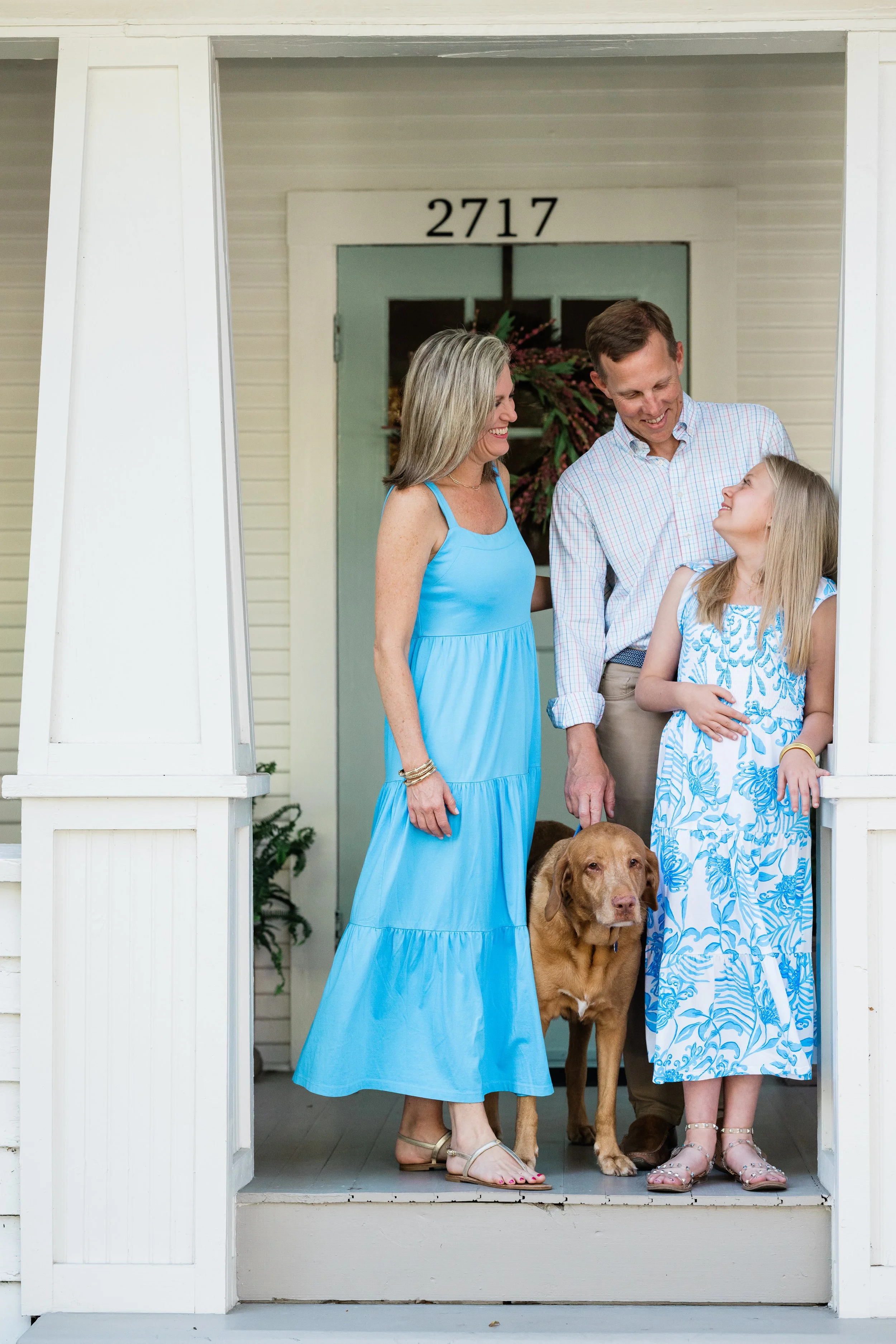 Family of three standing on porch with dog. Woman in blue dress, boy in checked shirt, girl in blue and white dress, house number 2717 behind them.