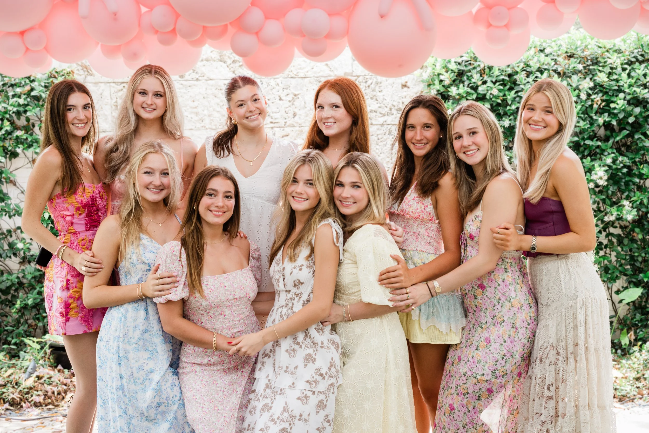 Group of teenage girls at a celebration, standing outdoors under pink balloons, wearing colorful summer dresses, smiling at the camera.