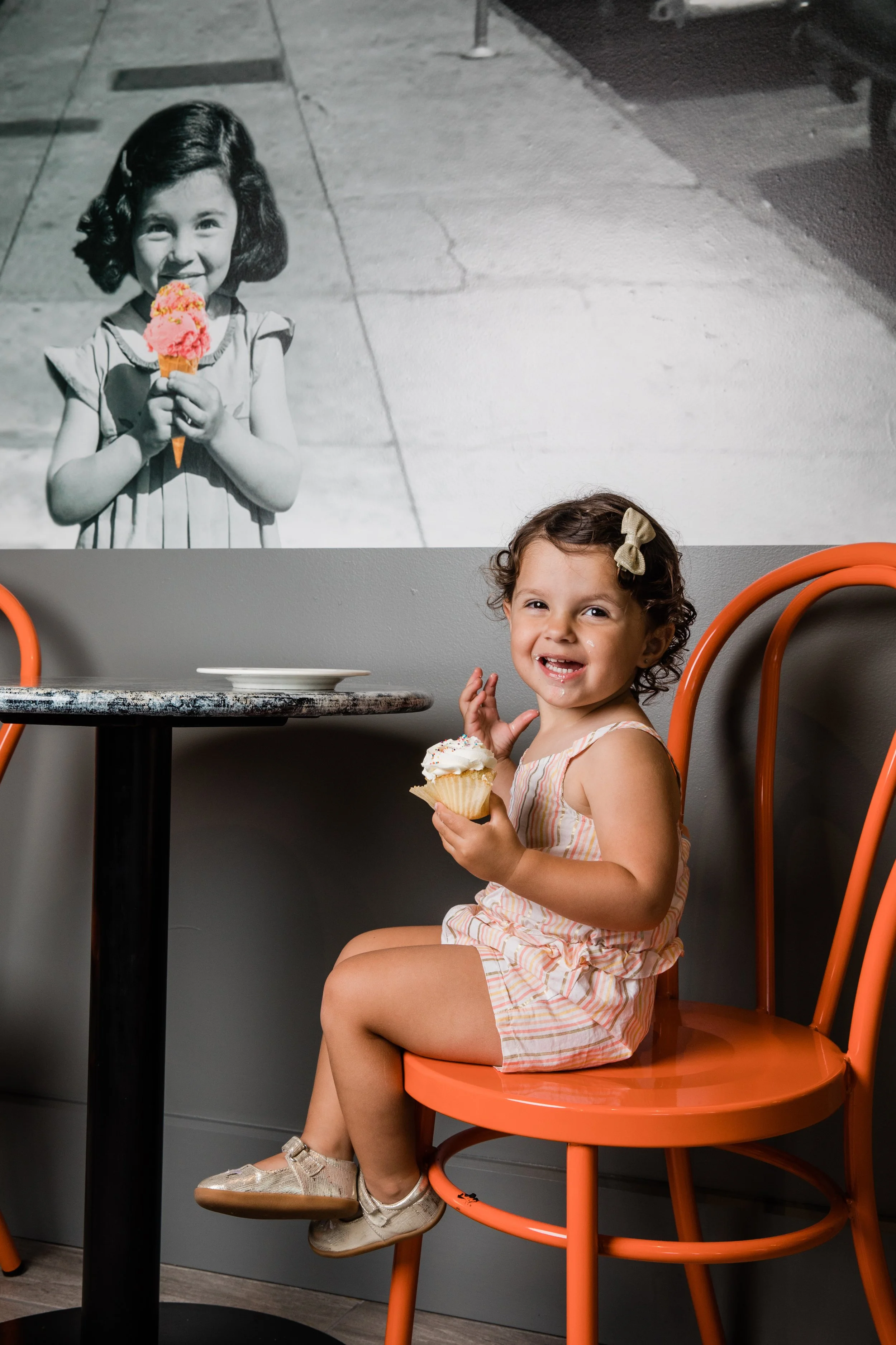 A laughing little girl perches on an orange chair at Little Midway ice cream shop. In the background, a vintage black and white mural shows a little girl that looks like her ancestor, holding an ice cream cone in selective color.