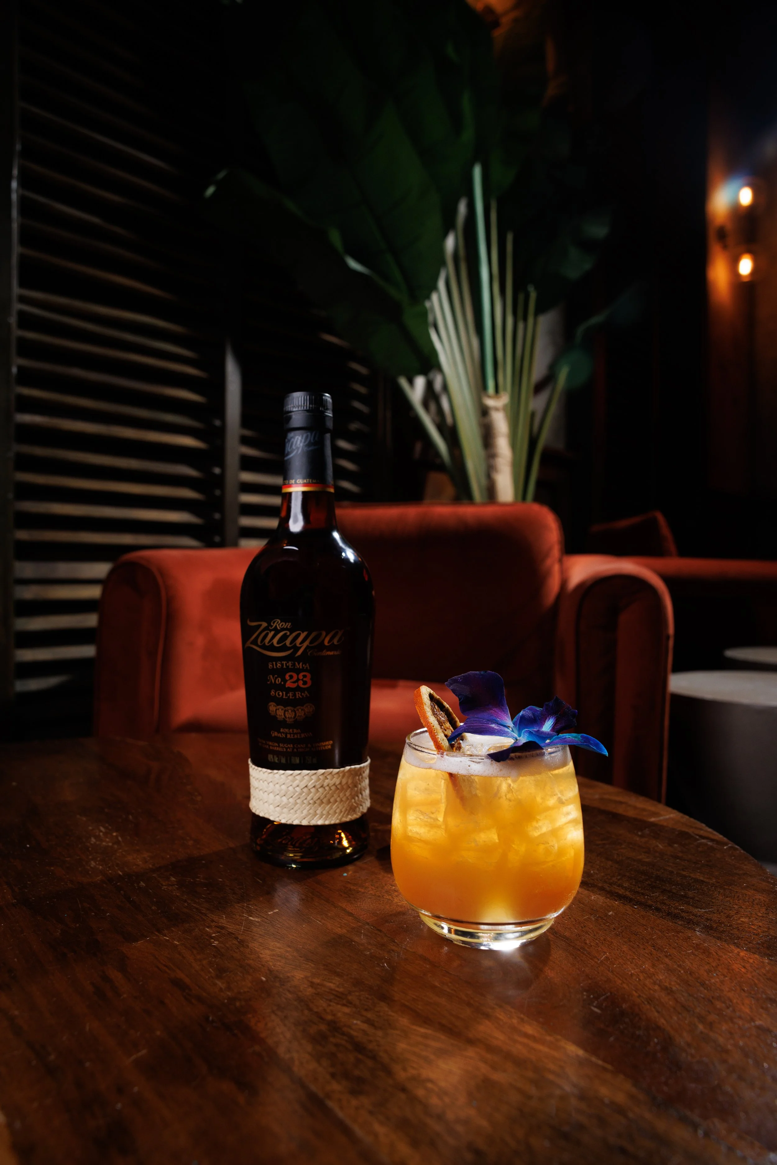 A bottle of Ron Zacapa rum, a cocktail garnished with edible flowers and a dried fruit slice, placed on a wooden table in a dimly lit lounge with a plant and wooden slats in the background.
