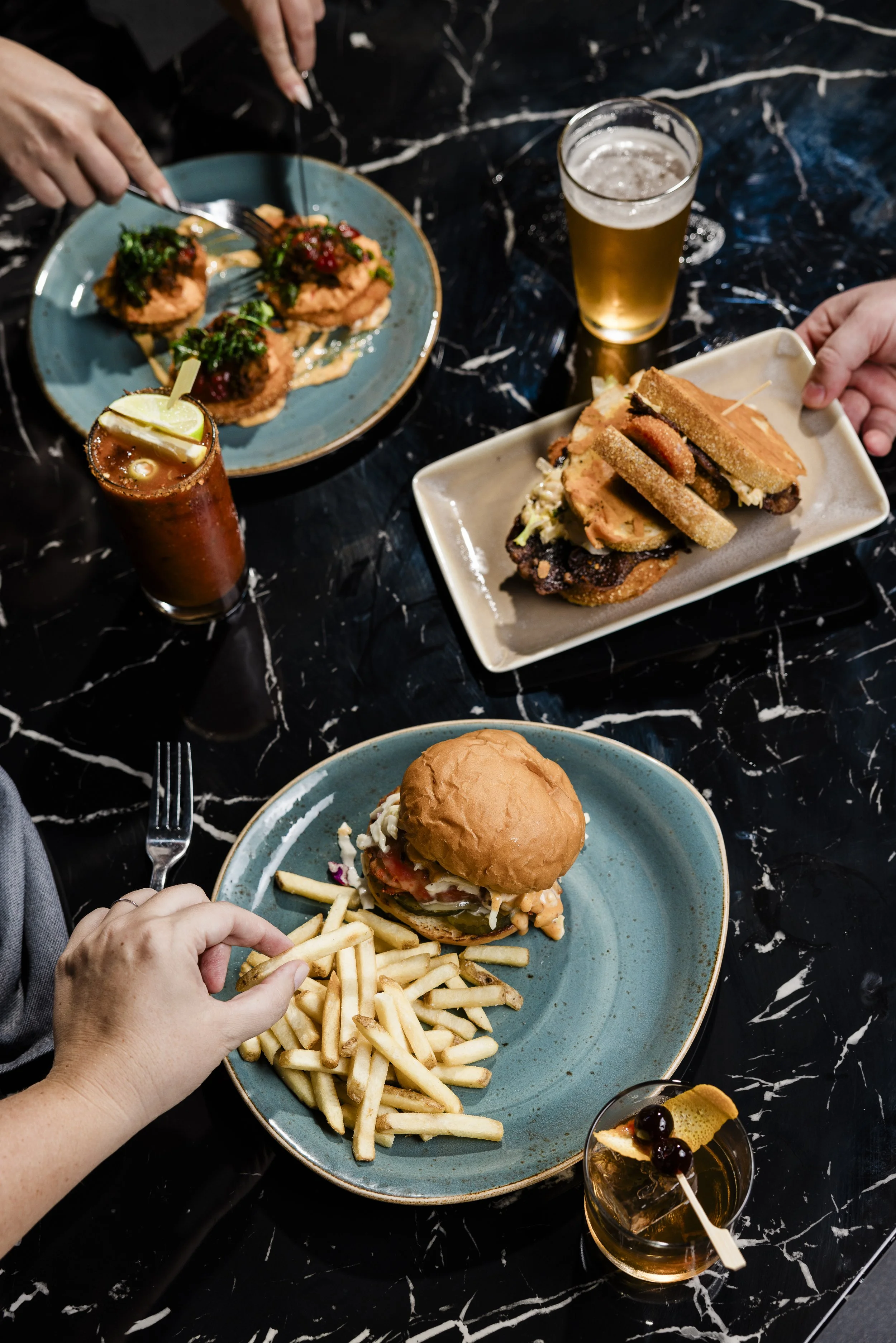 A table with various food and drinks including a burger with fries, a plate with fried chicken, and two glasses of beer, with hands reaching for the food.