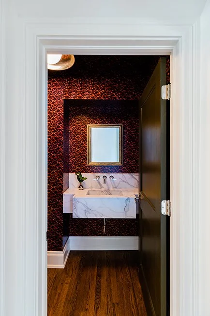 View through a bathroom doorway into a small space with dark patterned wallpaper, a white marble sink, a small mirror, and a vase with flowers.