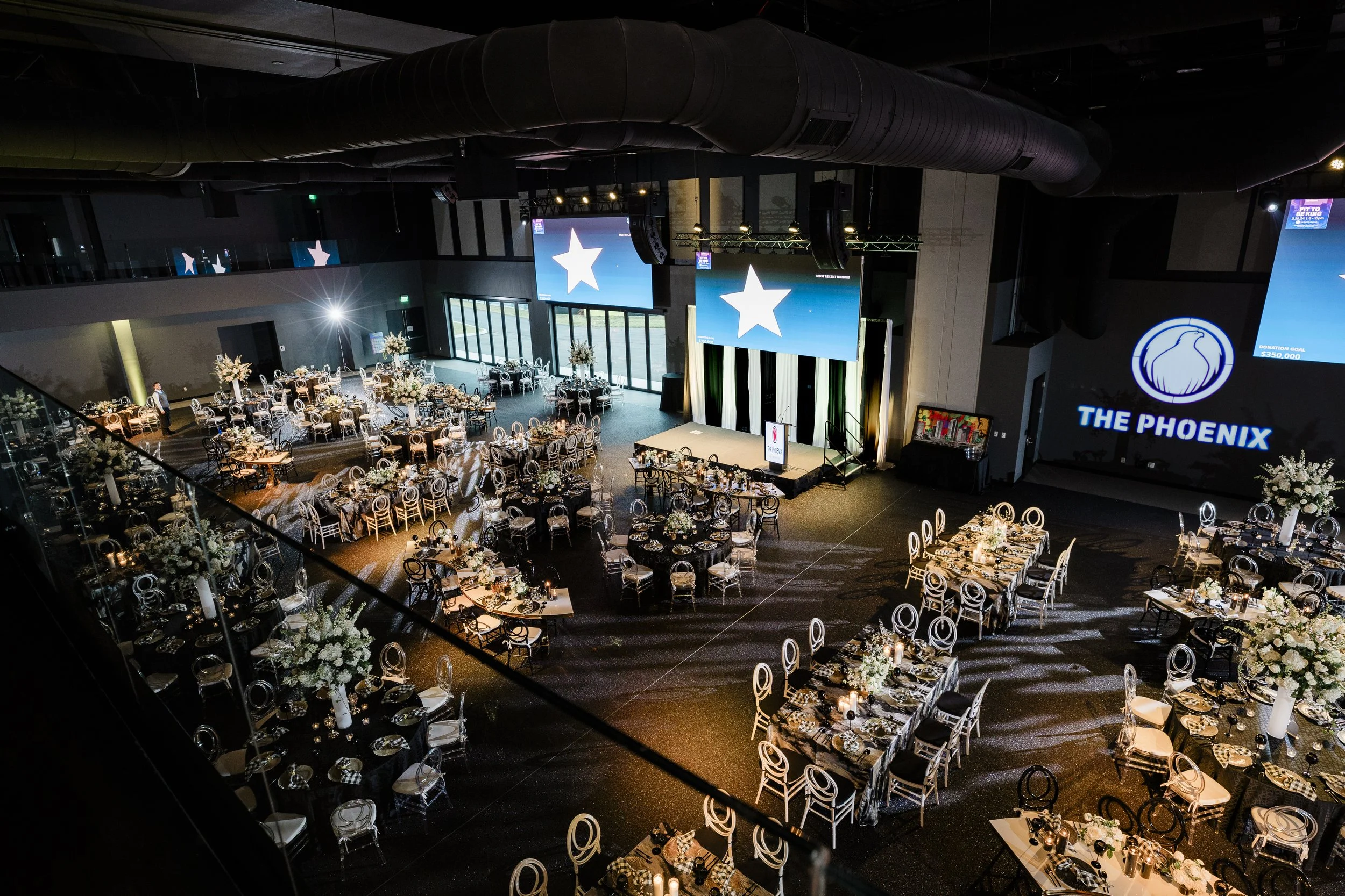 An event hall with decorated tables, floral centerpieces, and large screens displaying stars, set up for a formal celebration or gala at The Phoenix venue.