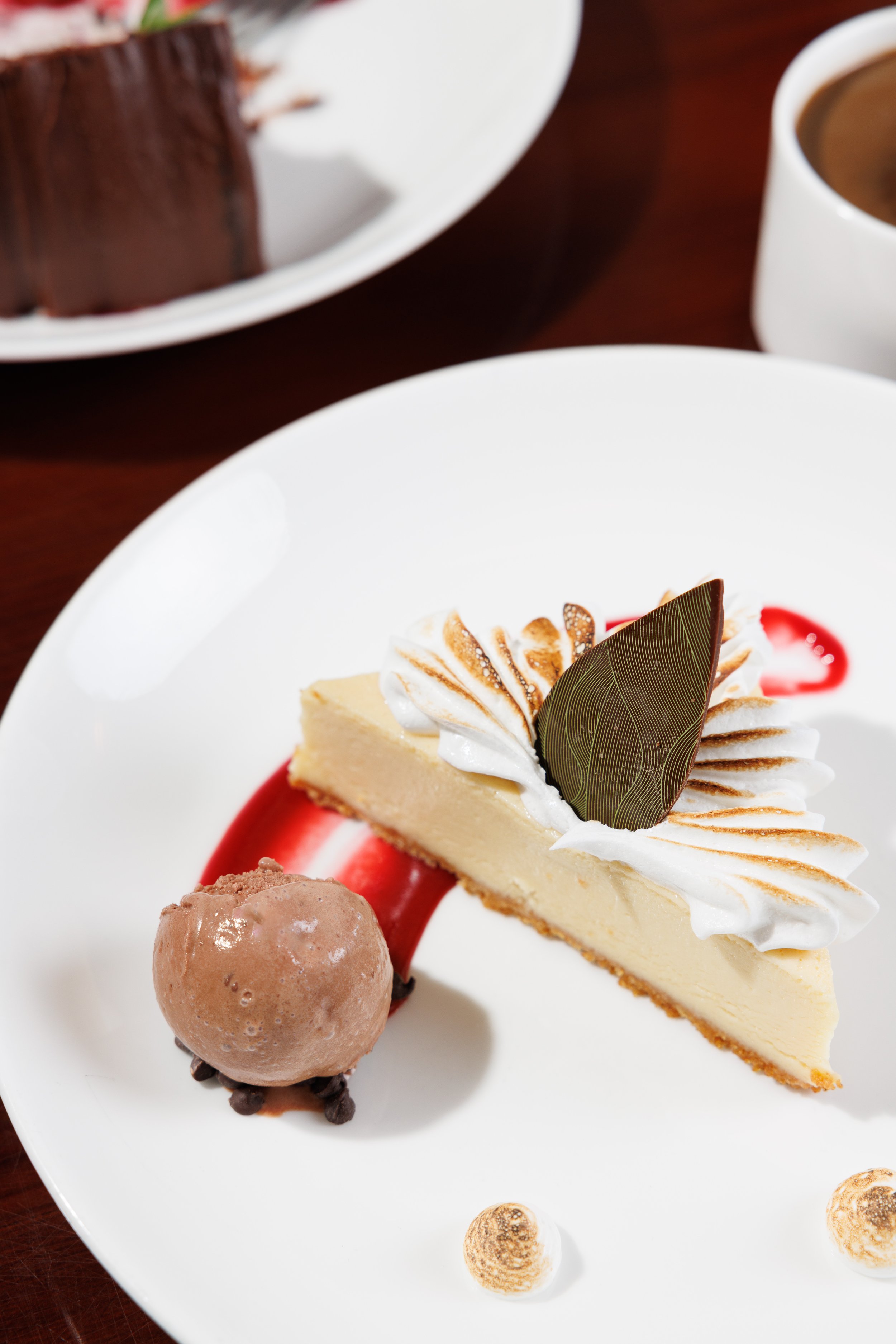 Slice of cheesecake with whipped cream, chocolate leaf, and red sauce on a white plate, alongside a scoop of chocolate ice cream with chocolate chips. A chocolate cake and coffee are visible in the background.