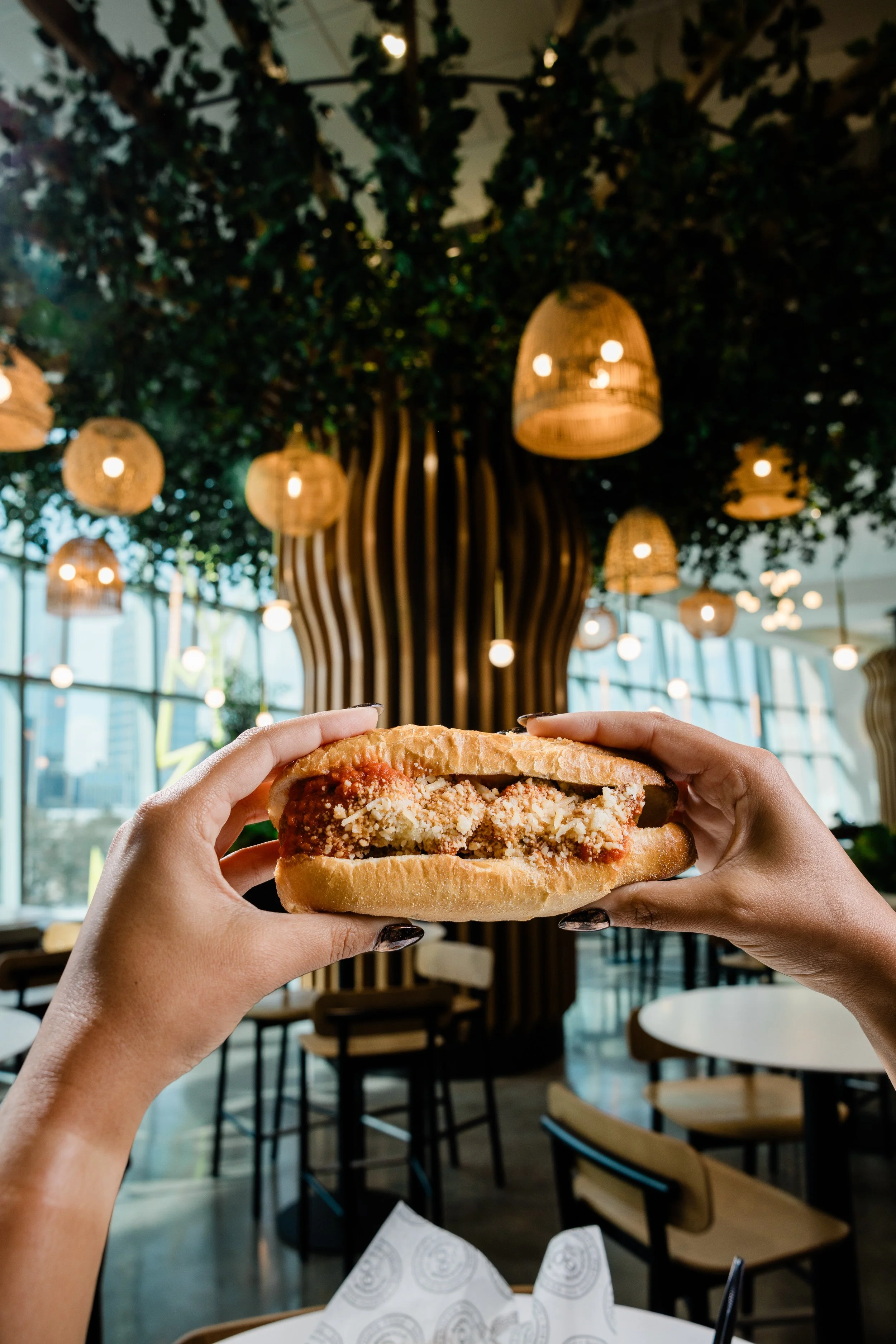 Hands holding a sandwich with breaded meat, cheese, and tomato sauce inside, in a restaurant with bamboo and leafy decor, hanging lamps, and large windows.