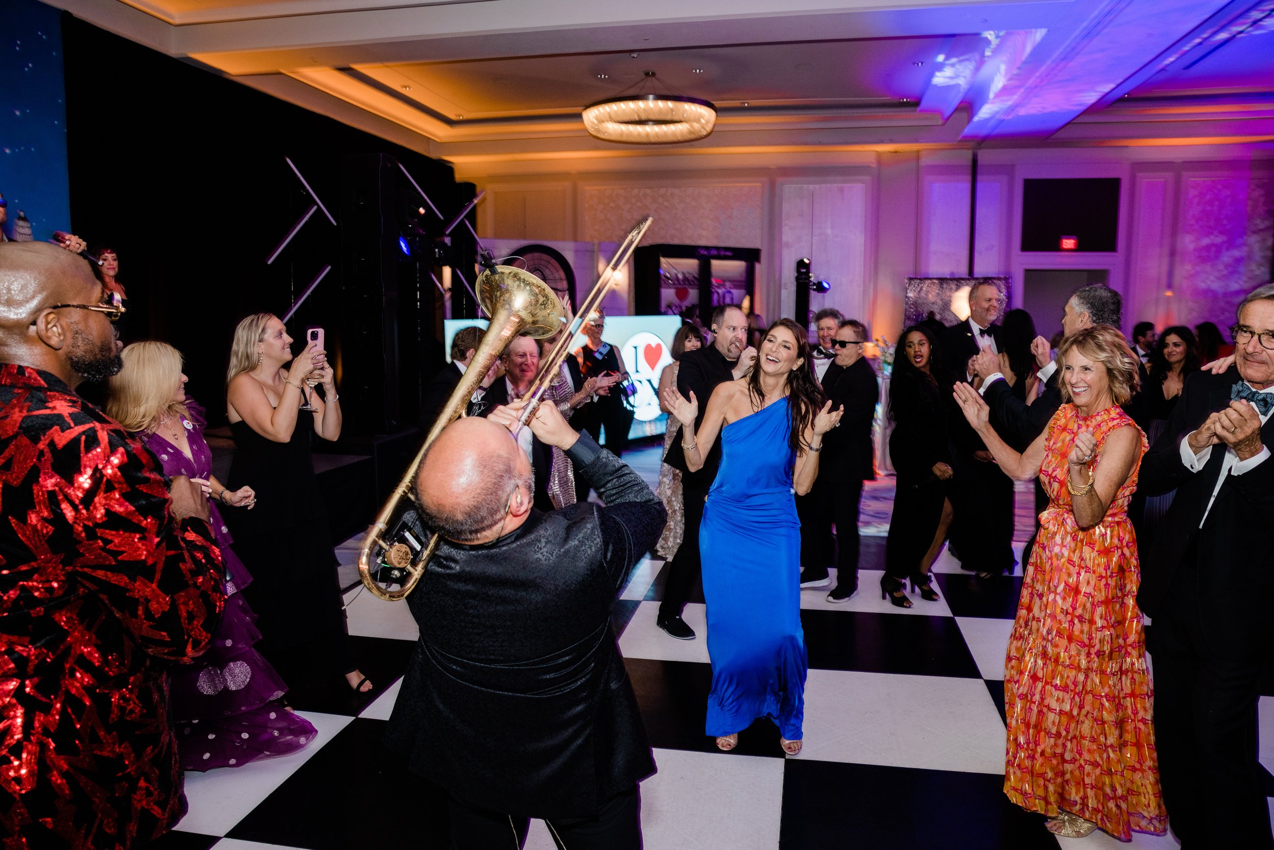 People dancing and celebrating at a lively party or event in an elegant ballroom with a chandelier. A woman in a blue dress and a man with a trombone are prominent among the cheerful crowd, some taking photos.