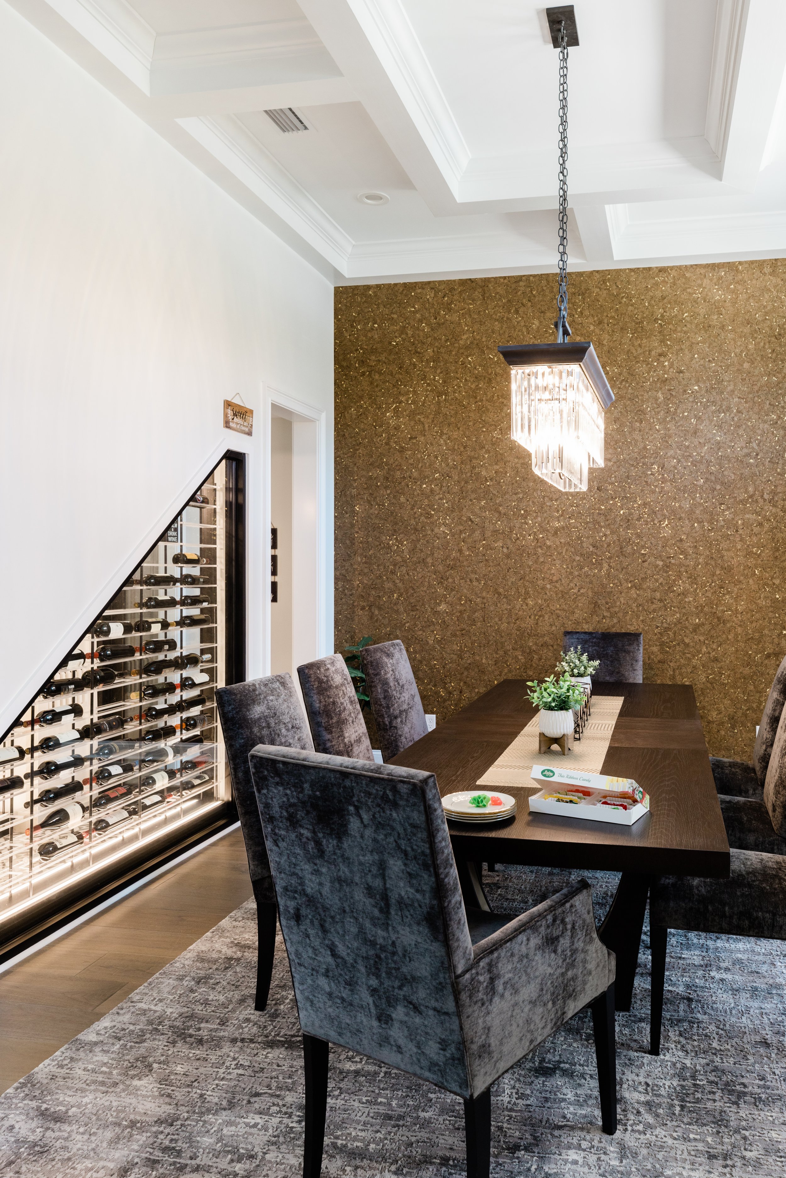 A dining room with a long dark wood table and six upholstered gray chairs. A small arrangement of potted plants and some plates are on the table. There is a textured gold accent wall and a white wall with a wine rack and a door. A modern chandelier h