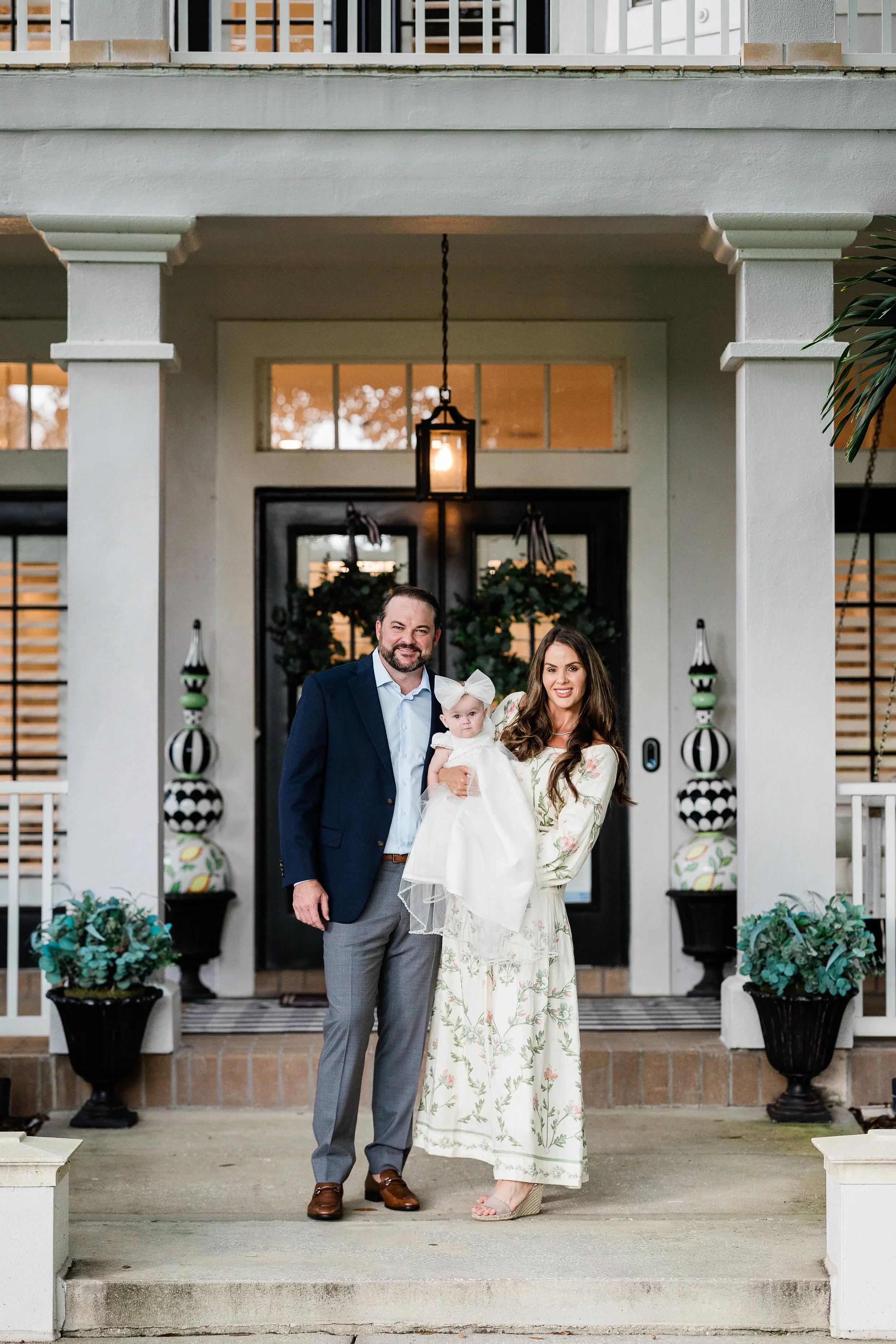 A family of three standing on the porch of a house, holding a baby girl dressed in a white dress with a large bow on her head. The man is wearing a navy blazer and gray pants, the woman is in a long floral dress, and they are smiling at the camera. T