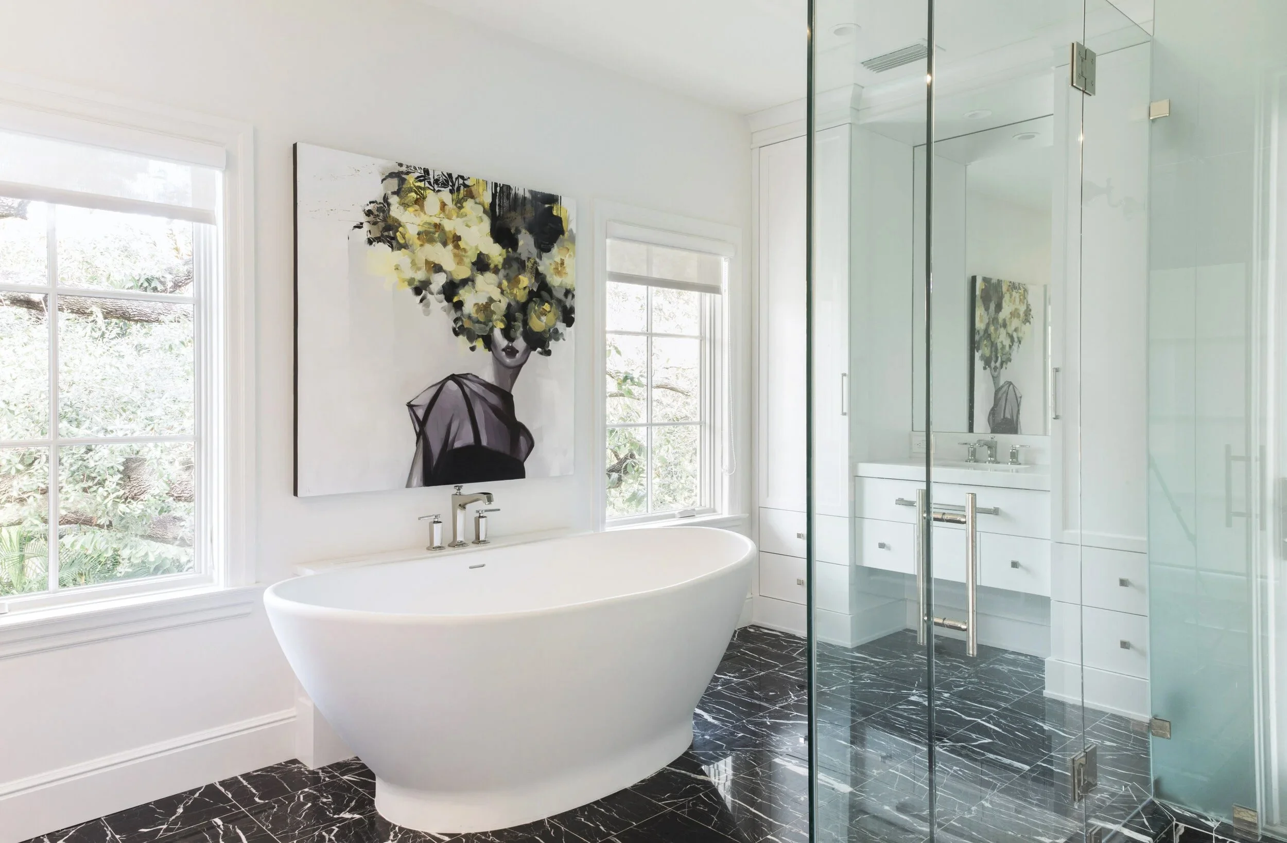 Modern bathroom with a white freestanding bathtub, black marble floor, and glass shower enclosure. Decor includes a colorful abstract portrait artwork on the wall, large windows with white blinds, and a mirrored cabinet above the sink.