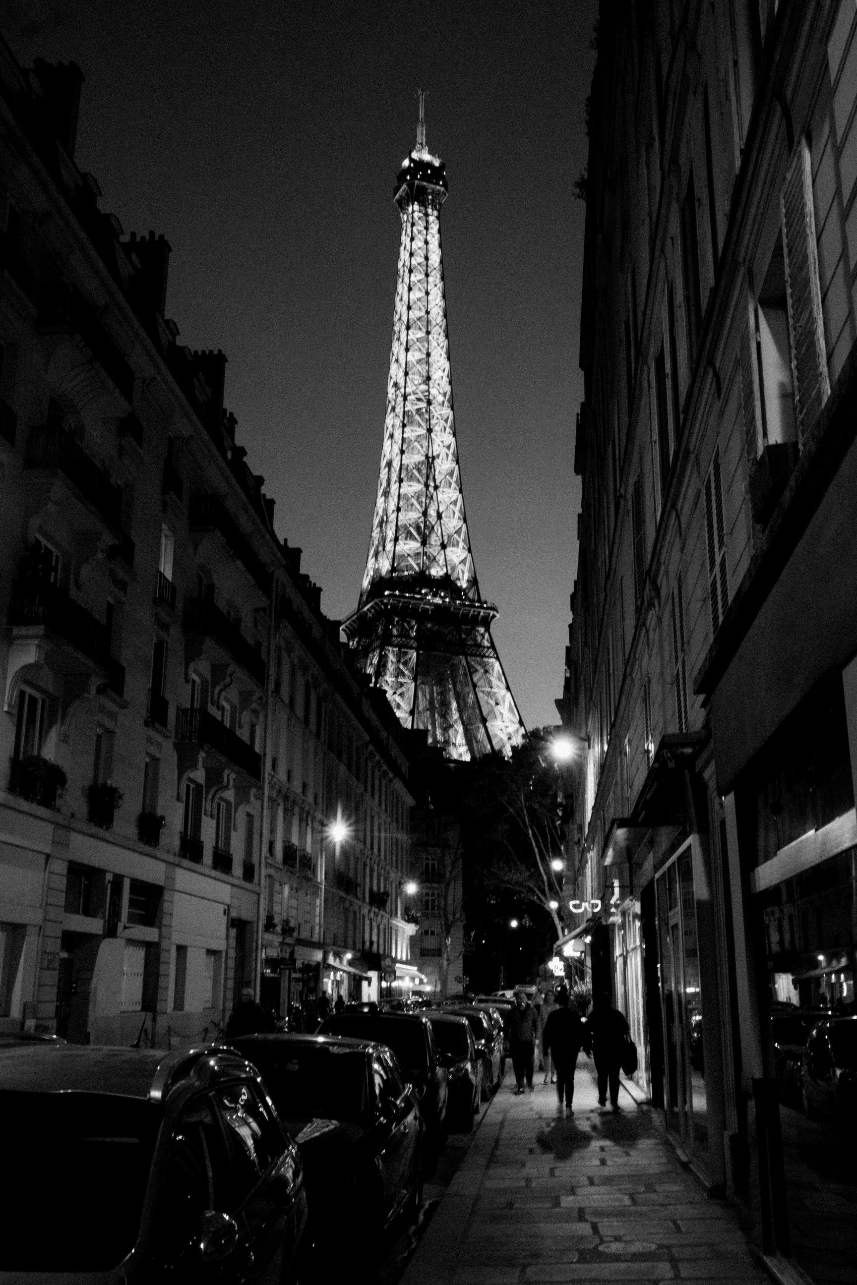 Nighttime street scene in Paris with the Eiffel Tower illuminated in the background. The street is lined with apartment buildings, cars parked along the curb, and a few pedestrians walking on the sidewalk.