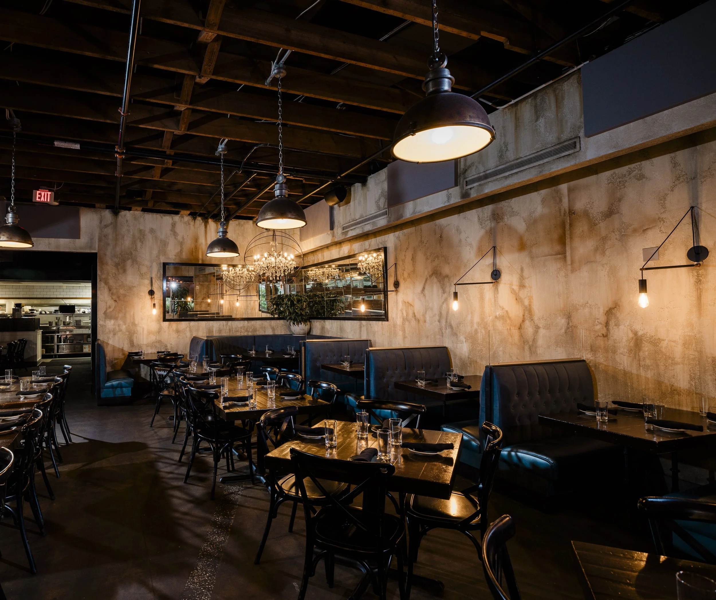 Interior of a restaurant with dim lighting, exposed wooden ceiling beams, hanging industrial-style pendant lights, a large mirror on the wall, and a chandelier. The dining tables are set with glasses and napkins, with black chairs and a dark wooden f