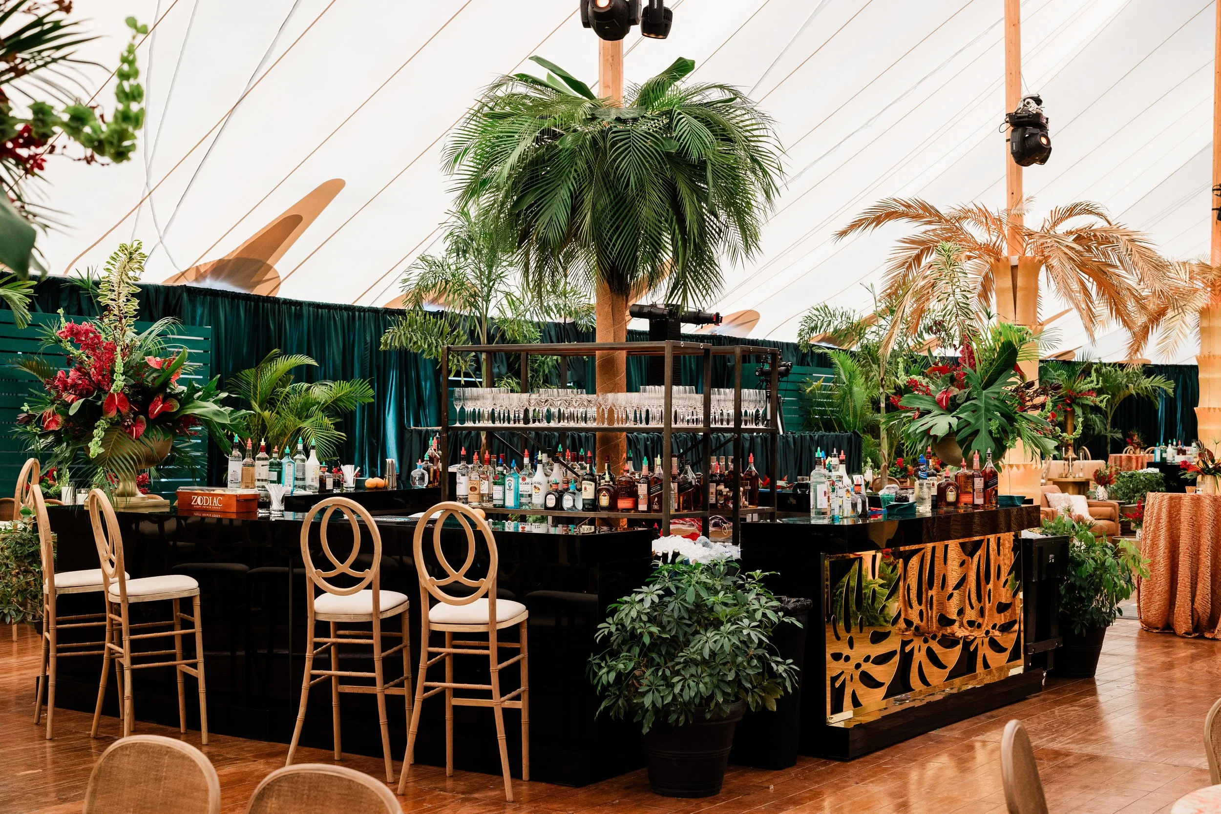 A party or event setup indoors with tropical-style plants, floral arrangements, a bar with various bottles of alcohol, glasses, and bar tools. There are chairs around the area and lighting fixtures hanging from the ceiling.