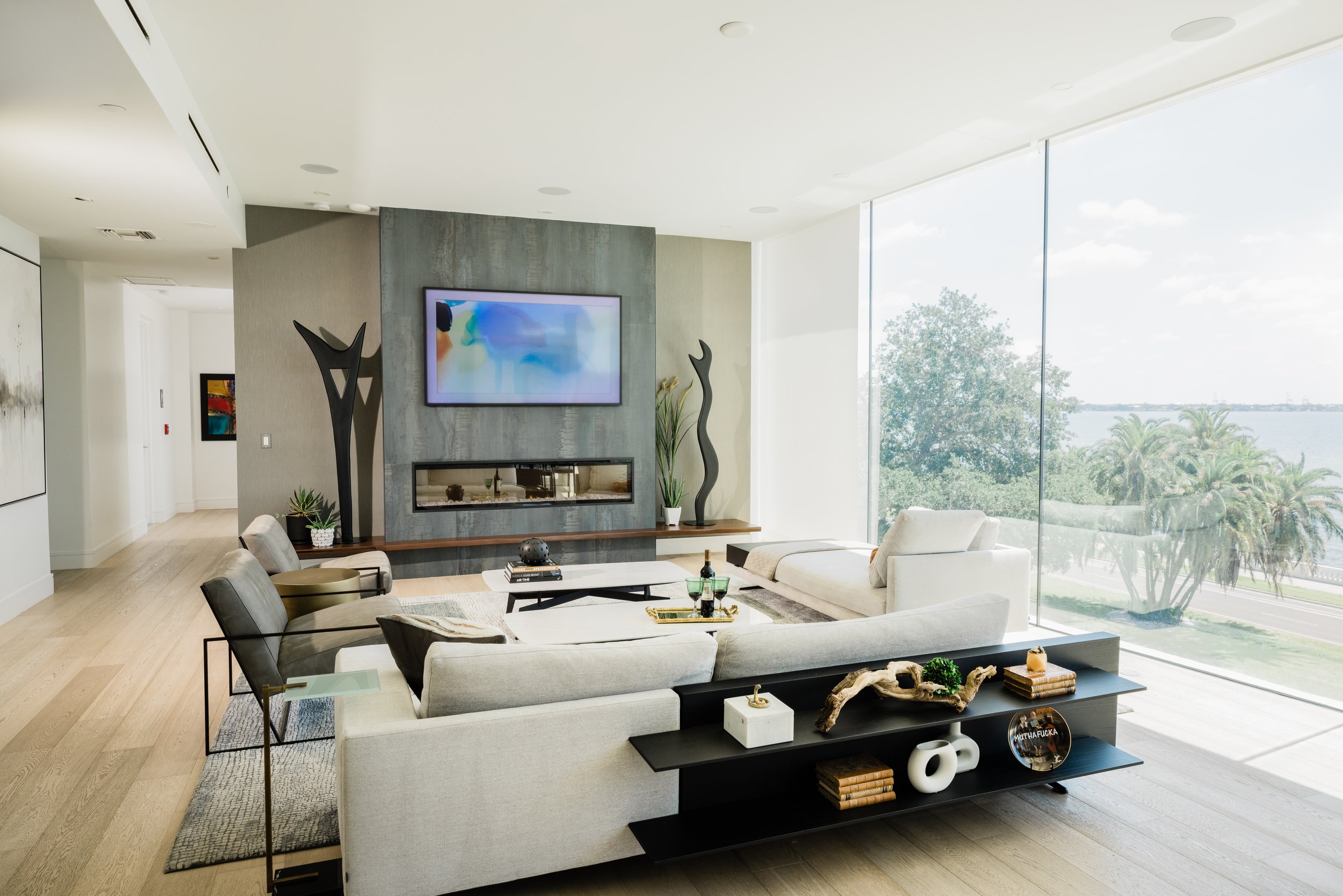 A modern living room with large floor-to-ceiling windows overlooking a scenic water view. The room features a light-colored sectional sofa, a black coffee table, a TV mounted on a textured gray wall, and contemporary decor including plants, sculpture