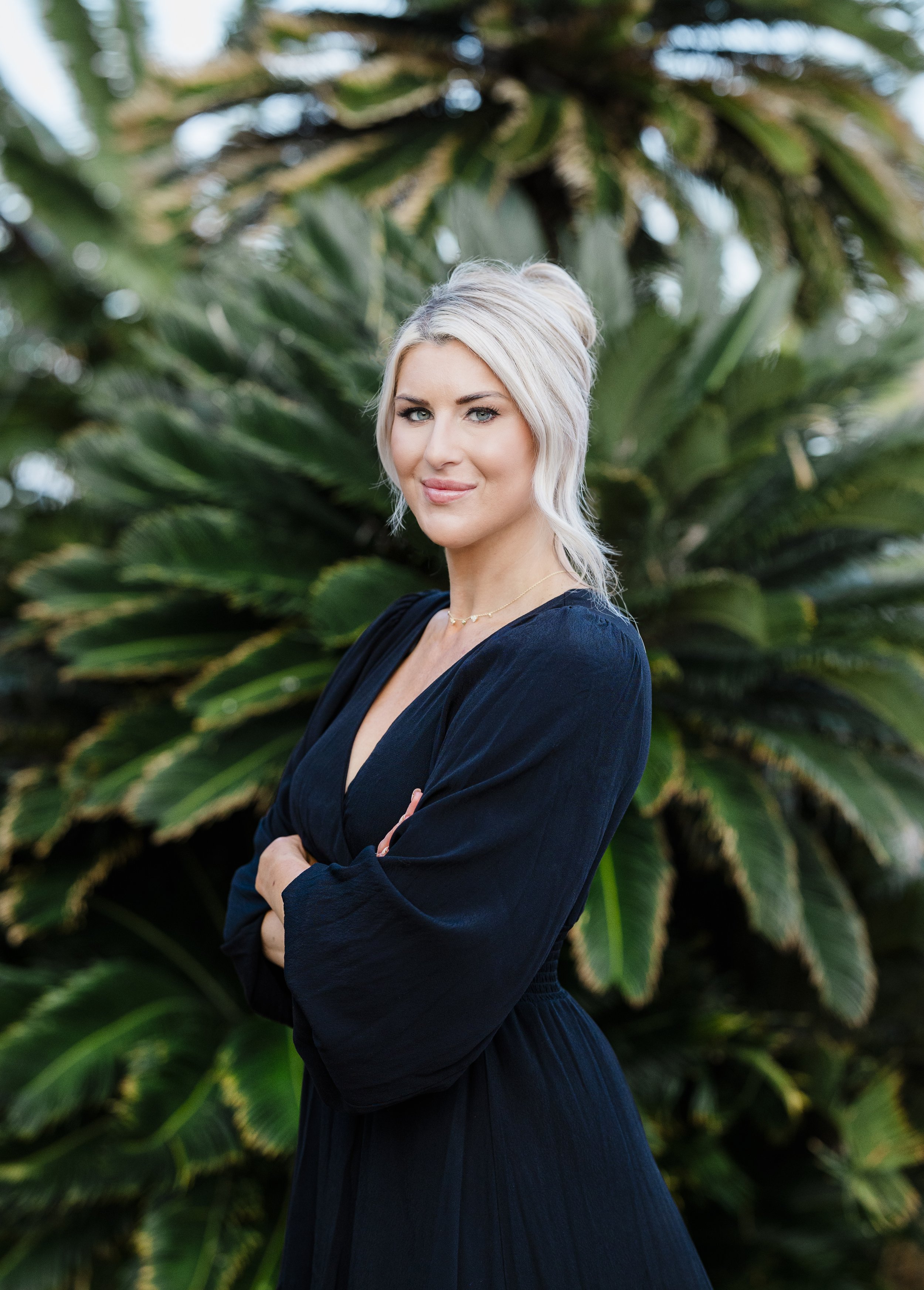 A woman with platinum blonde hair styled in a loose bun, standing outdoors with her arms crossed, in front of green tropical plants with large leaves.