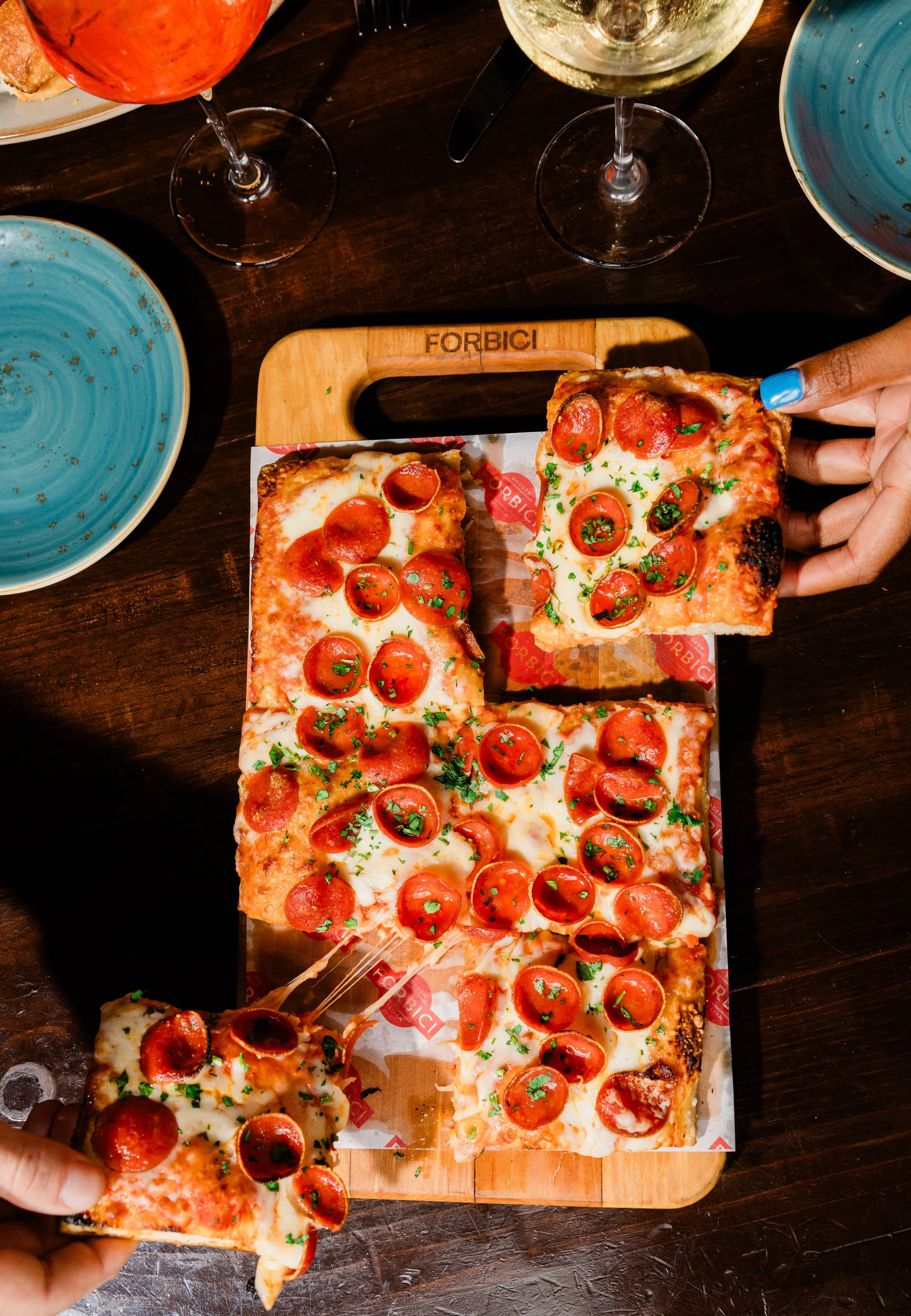 A large pepperoni pizza on a wooden serving board with a person's hand holding a slice. The pizza has cheese, pepperoni, and green herbs. Surrounding the pizza are empty plates, glasses of white and pink wine, and part of a plate with food on a dark 